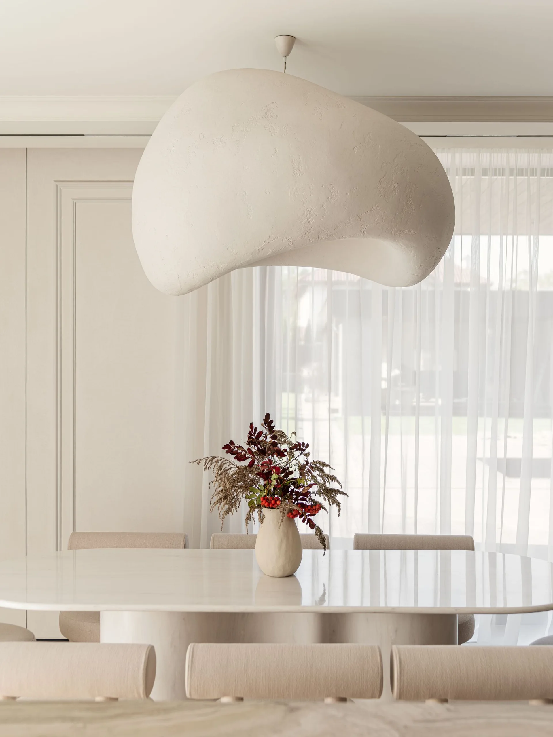 A white oval dining table with beige chairs sits beneath a large, organic-shaped white pendant light in a minimalist room with sheer curtains.