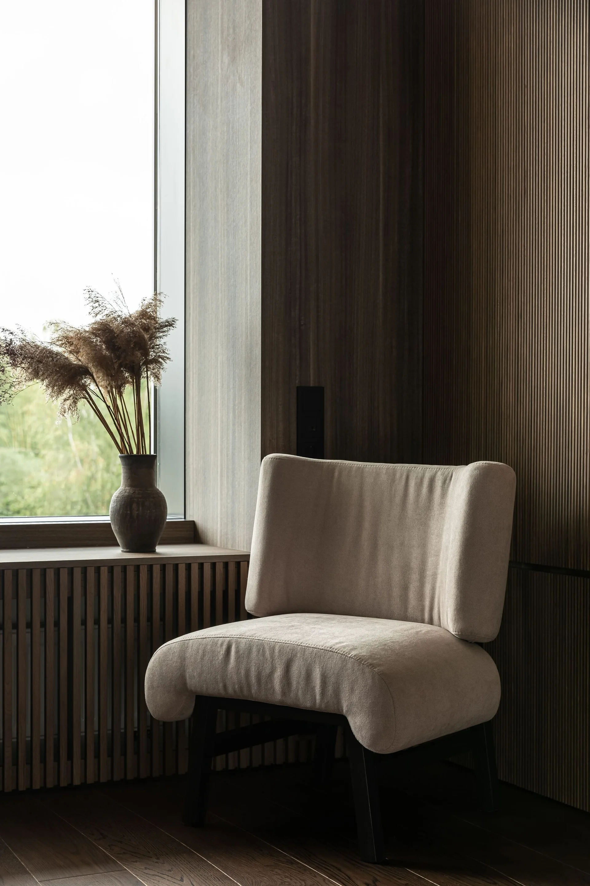 A cream upholstered armchair sits beside a window with a slatted wood cover, against dark wood-paneled walls.