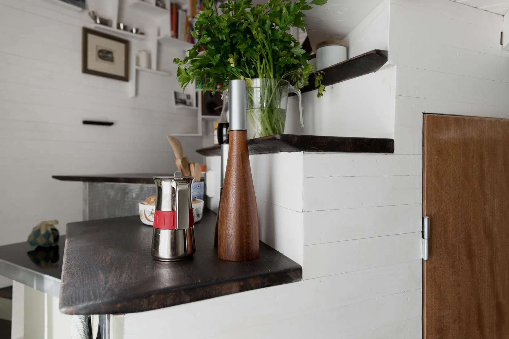 A close-up of a dark stained wood kitchen counter with a metal moka pot and wooden pepper mill against a white paneled wall.