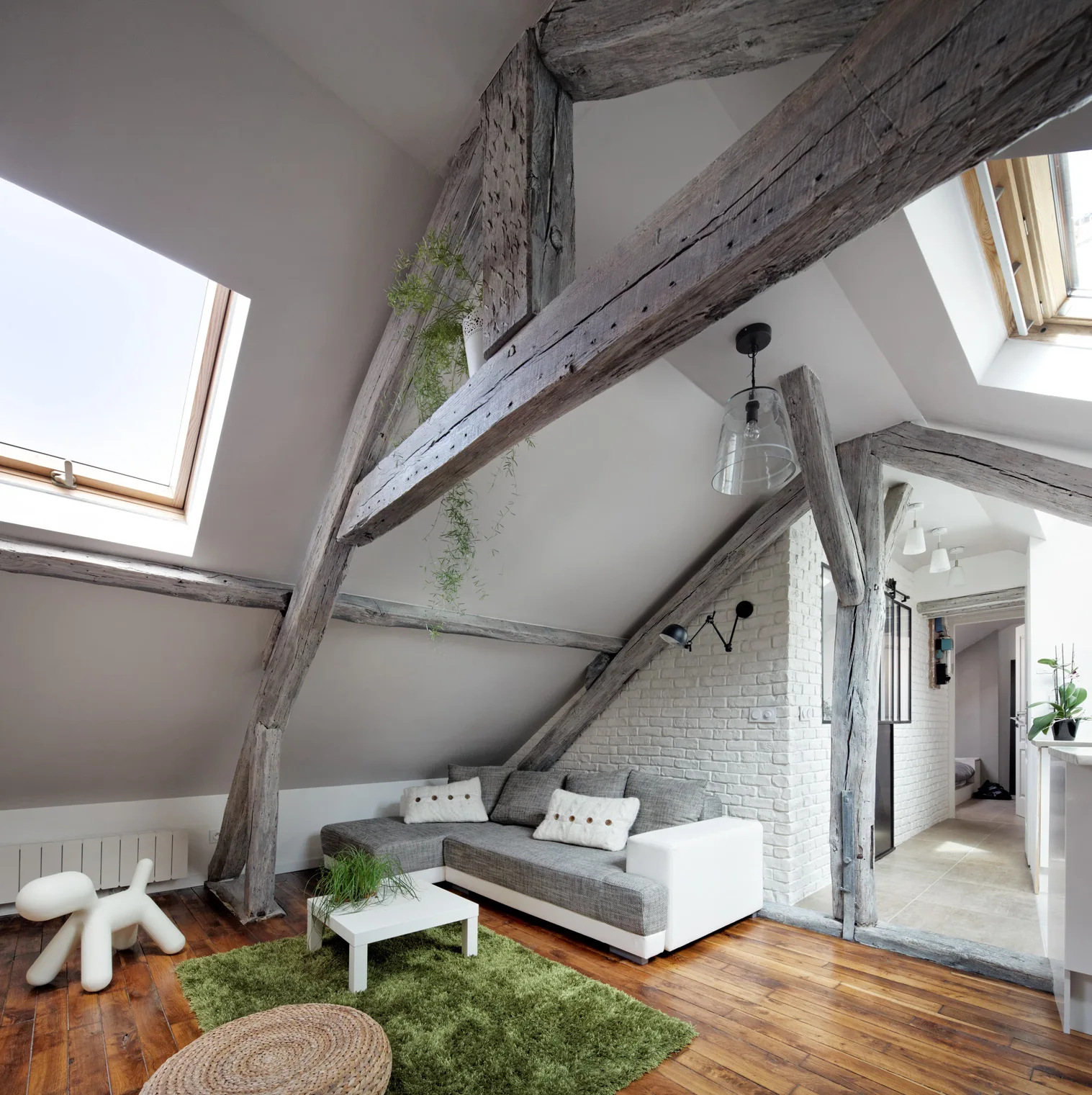 An attic living room featuring a grey sectional sofa, a green rug, and rustic exposed wooden beams under a sloped ceiling with a skylight.