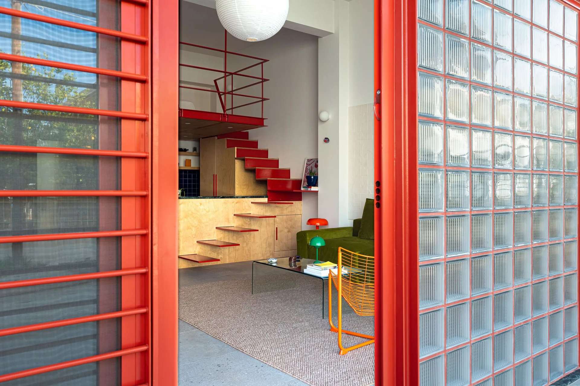 A view through a red doorway into a modern loft living room featuring a green sofa, glass coffee table, and a plywood storage unit with a red staircase.