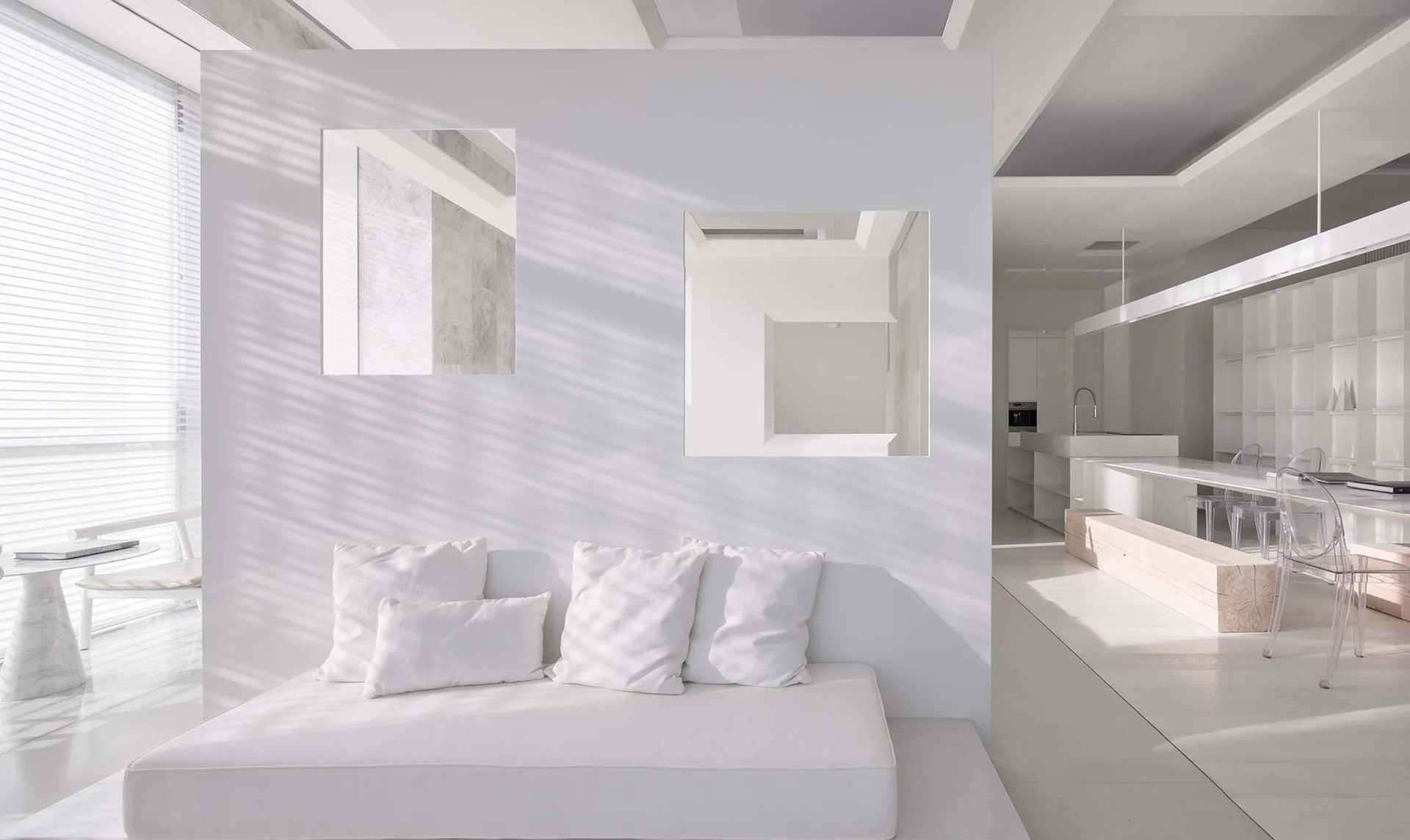 A minimalist white living room features a low bench sofa set against a central partition wall with two square cutouts.