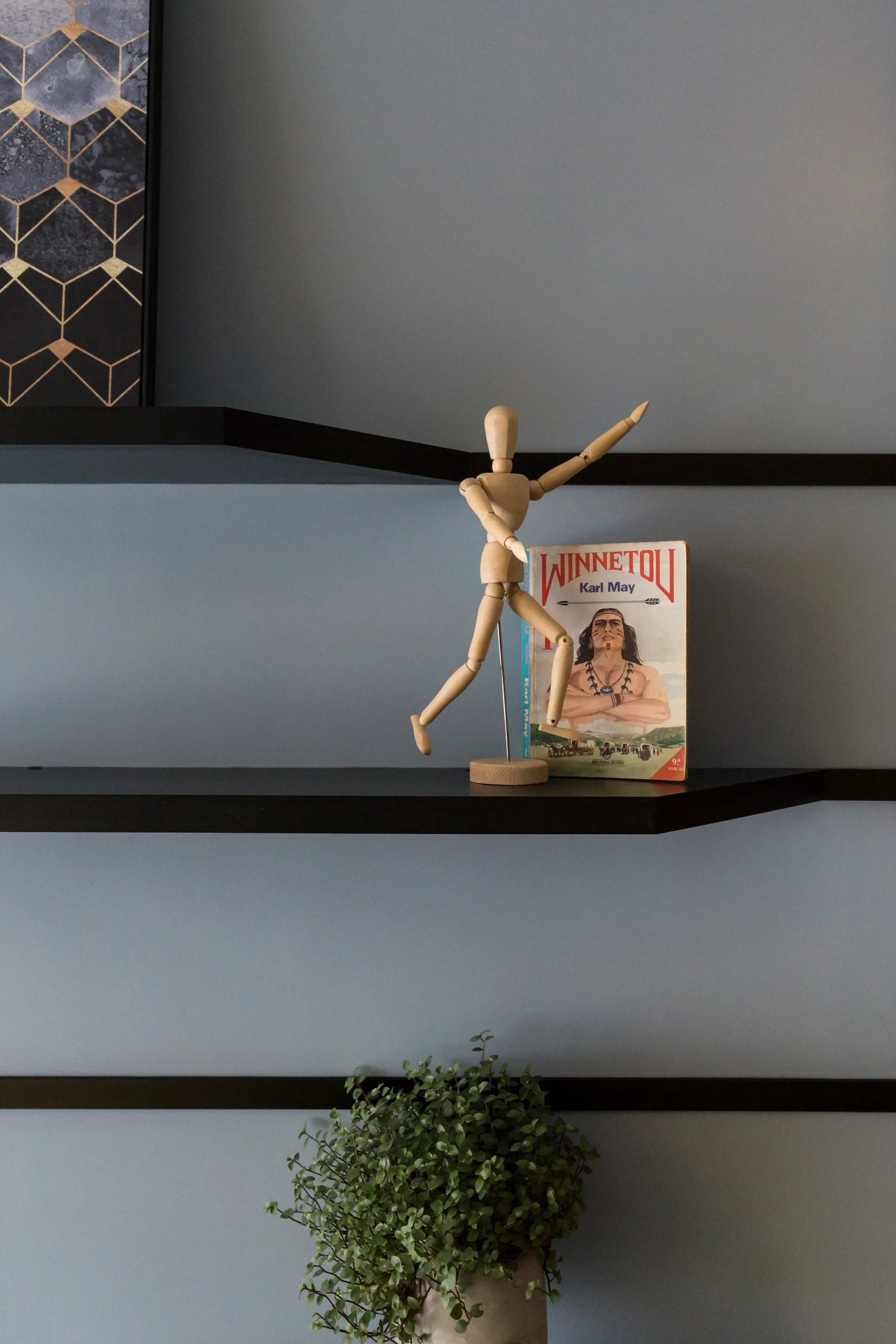 Black floating shelves on a matte gray wall displaying a wooden artist mannequin, an illustrated book, and a green potted plant.
