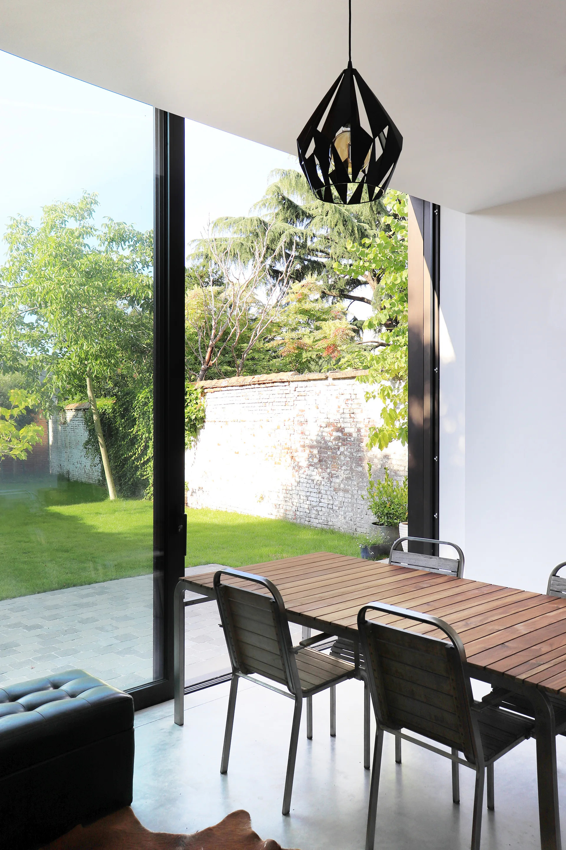 A modern dining room features a wood slatted table with metal chairs next to a large sliding glass door opening to a grassy yard.