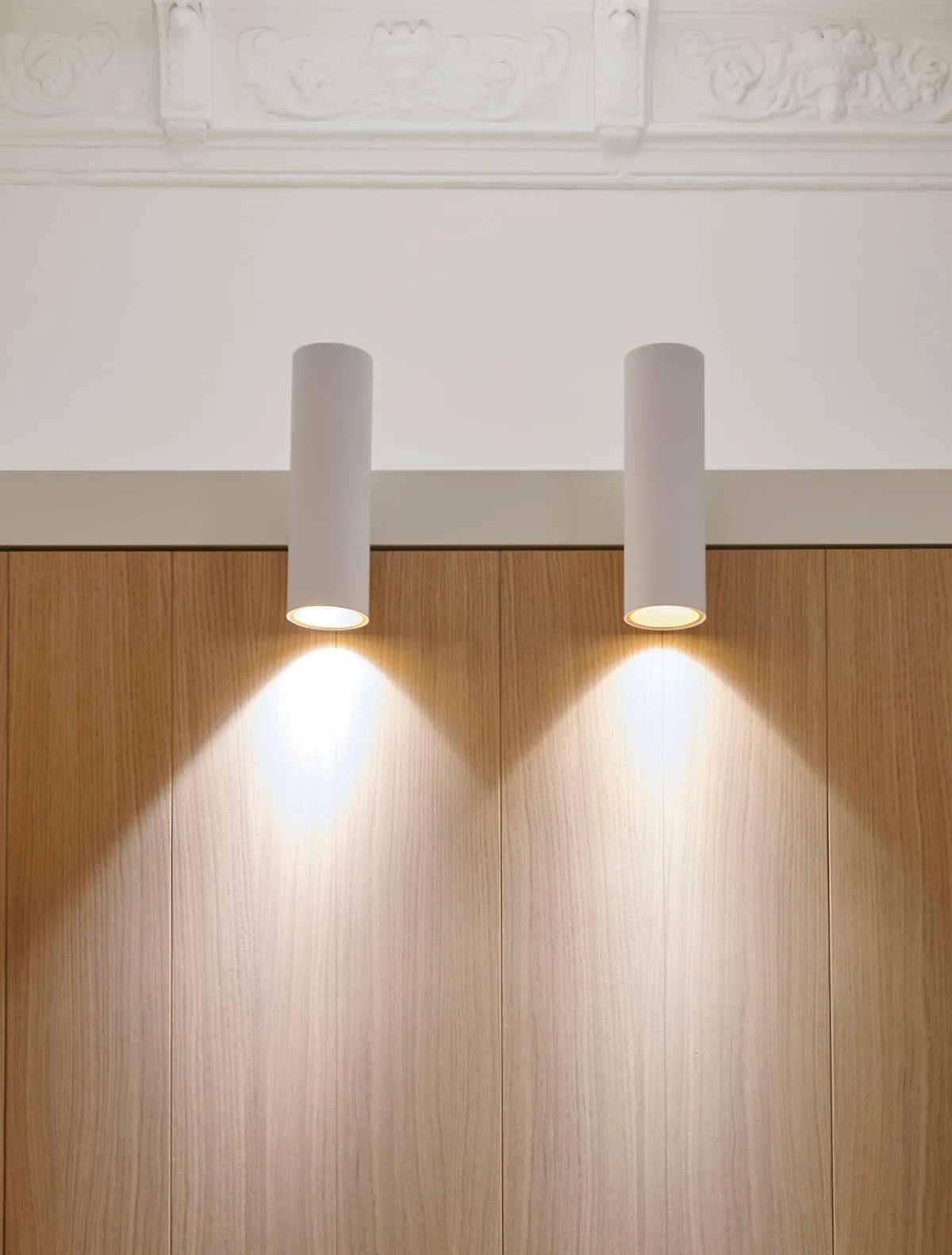 Two white cylindrical spotlights mounted on a light wood wall below ornate ceiling molding, casting warm light downward.