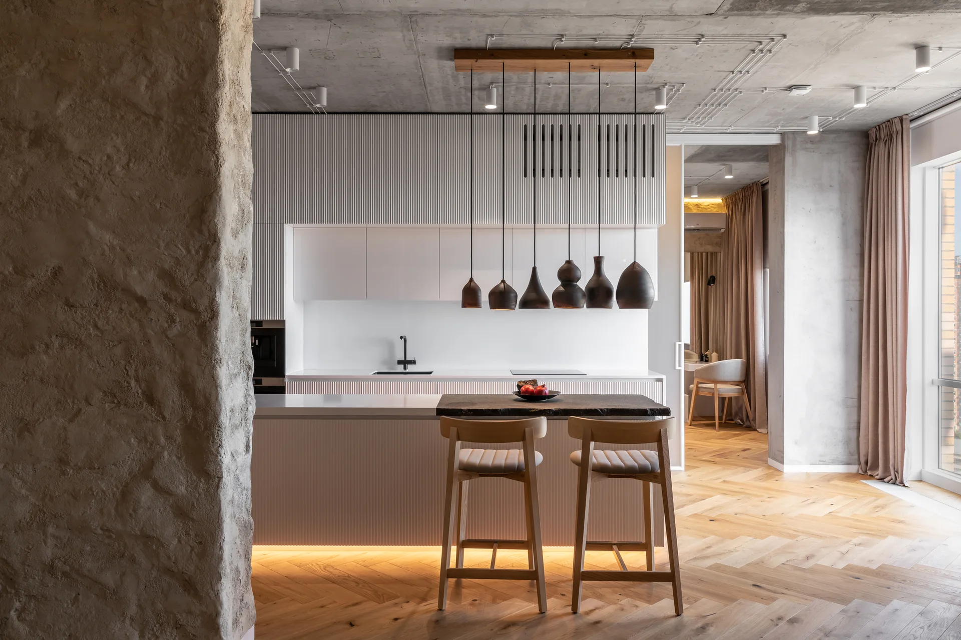 A modern kitchen featuring a white fluted island with wooden stools, a cluster of dark pendant lights, and an exposed concrete ceiling.