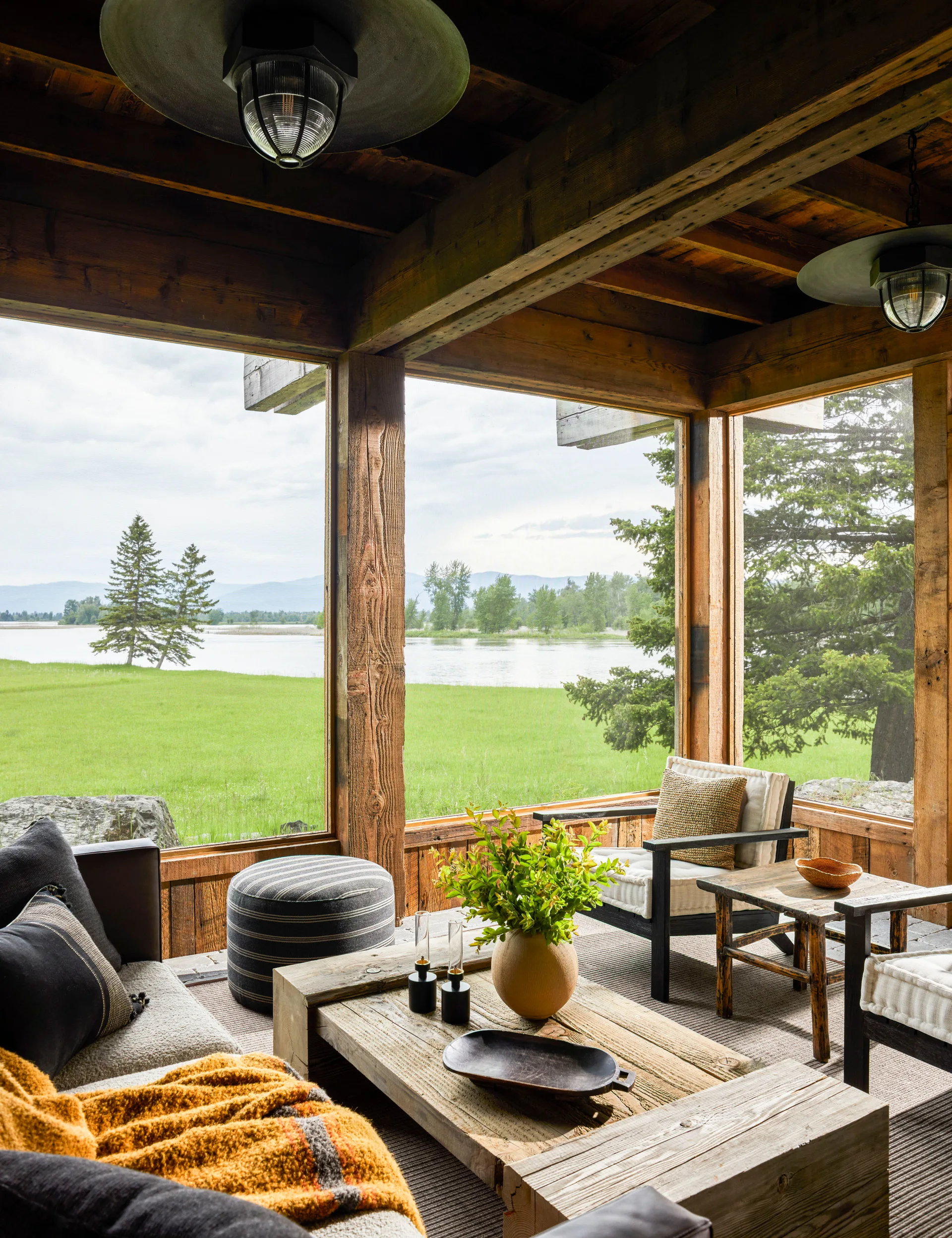 A rustic screened-in porch features heavy timber framing, a low wooden coffee table, comfortable outdoor furniture, and views of a grassy field and lake.
