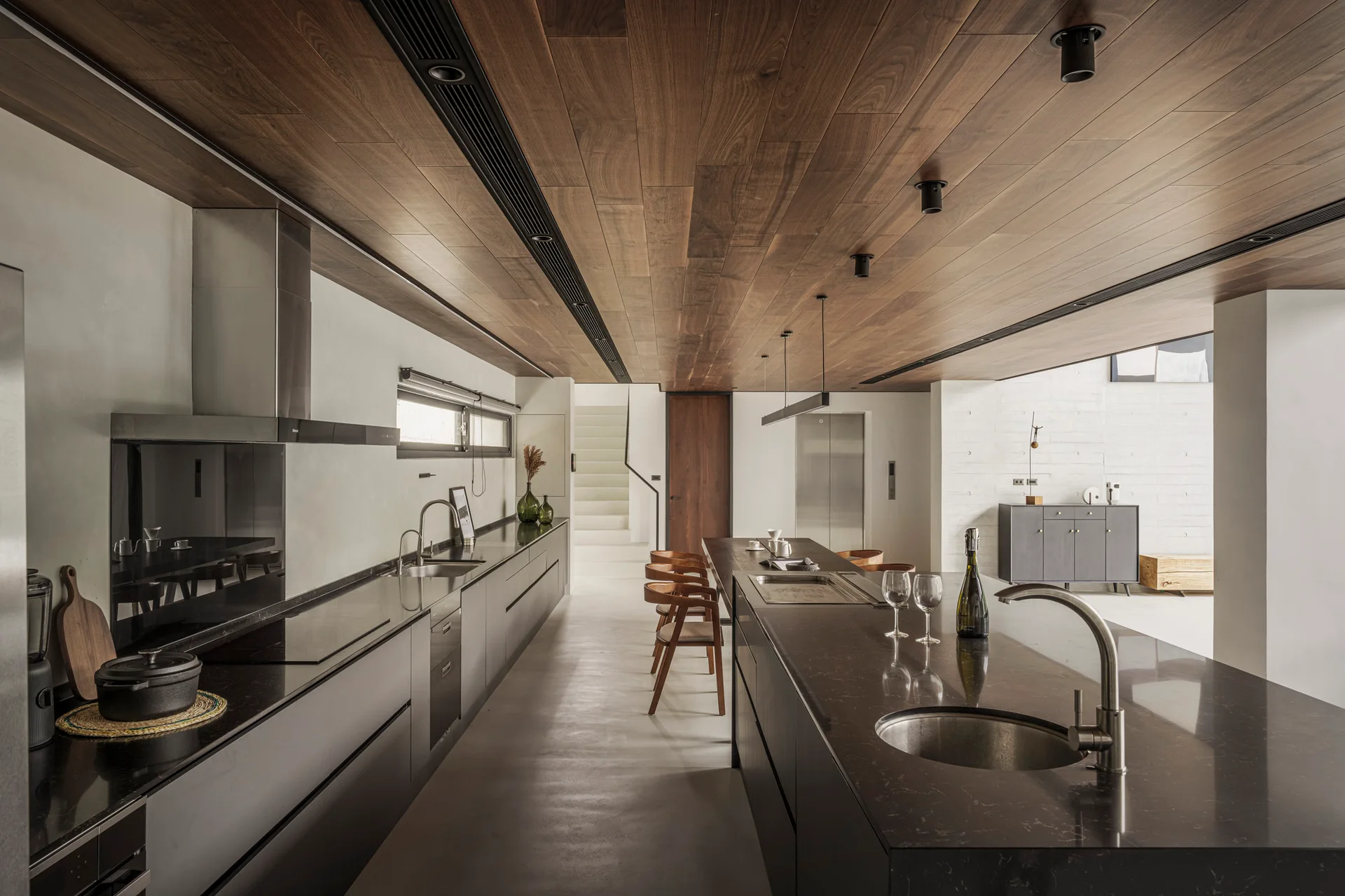 A minimalist kitchen features a long dark stone island and matching charcoal cabinetry beneath a rich wood plank ceiling with recessed lighting.