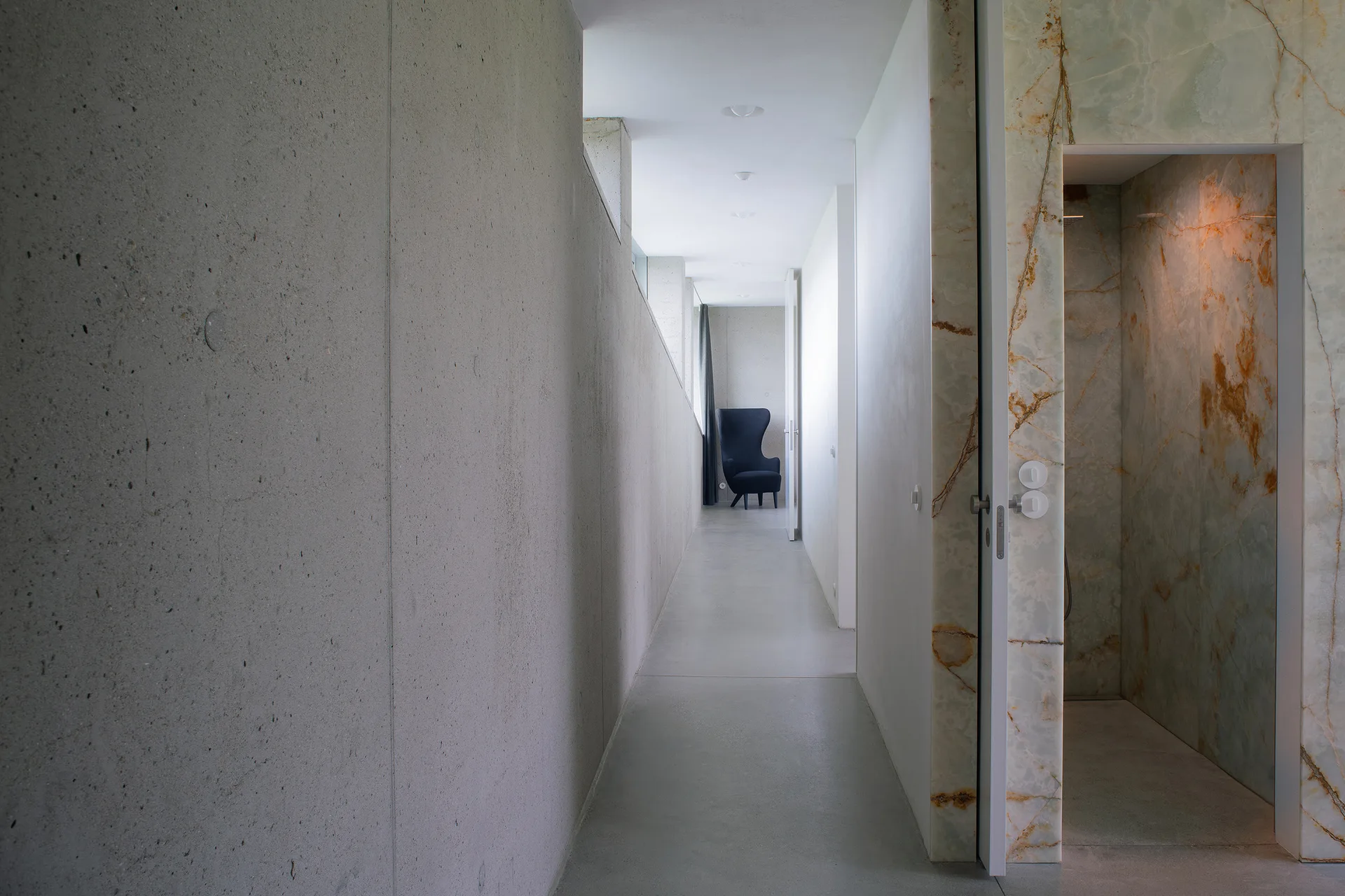 A minimalist concrete hallway leading to a distant dark armchair features a polished, veined stone doorway cladding on the right.