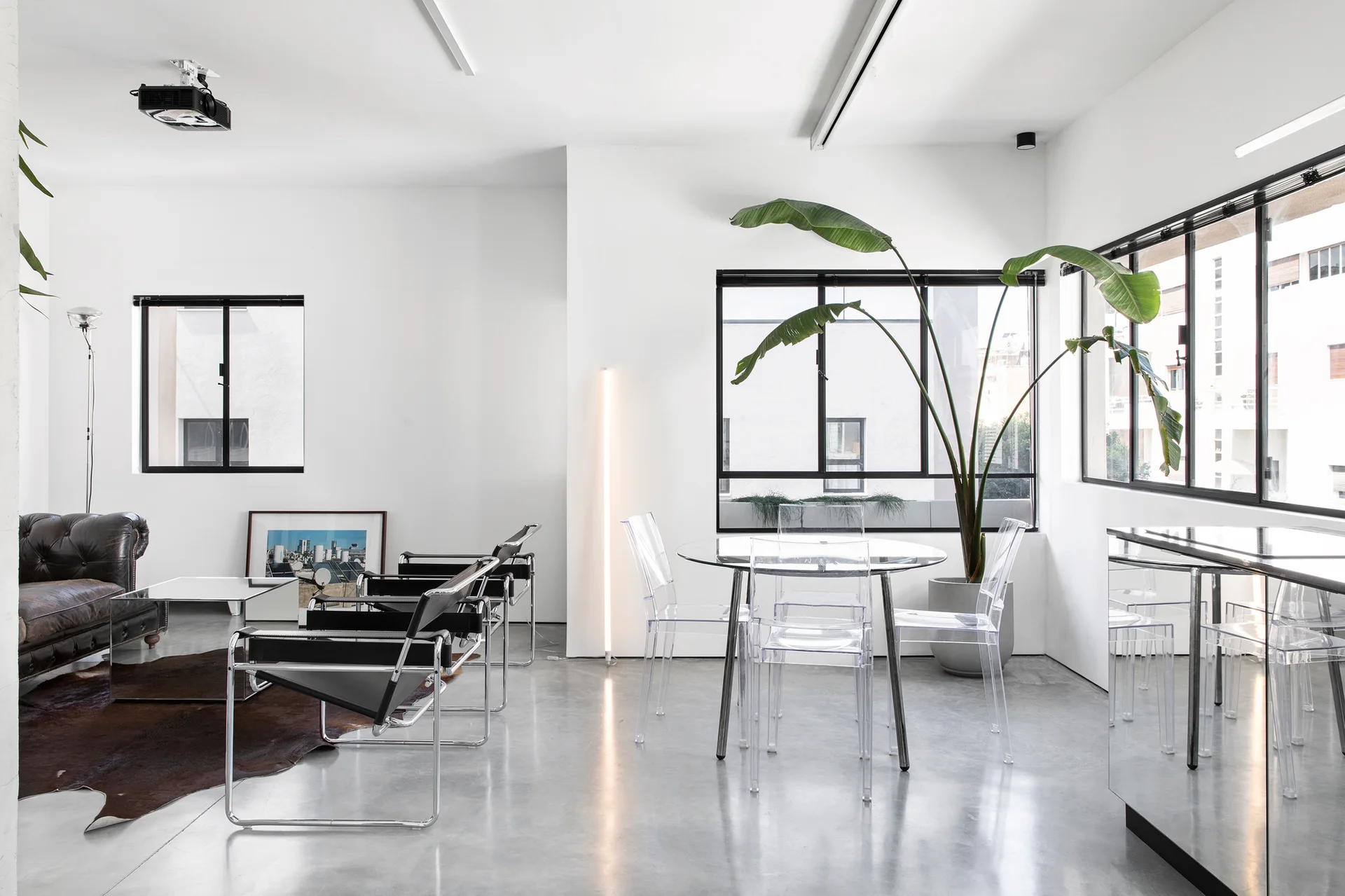 An open living space with polished concrete floors, featuring a black leather sofa, chrome chairs, and a round glass dining table with acrylic chairs.