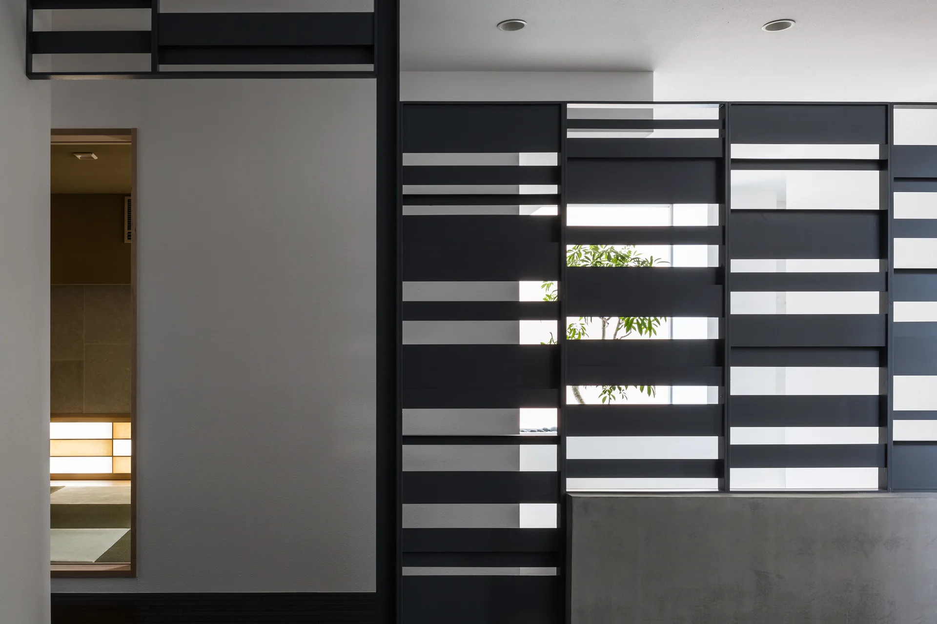 A matte black metal slatted screen sits on a concrete base next to a white wall, revealing a green plant and a doorway to another room.