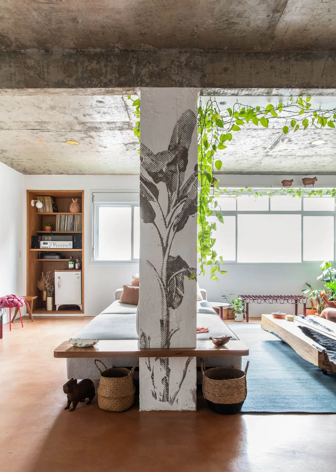 A loft living room featuring an exposed concrete ceiling, a grey sofa, and a central structural pillar decorated with a botanical illustration.
