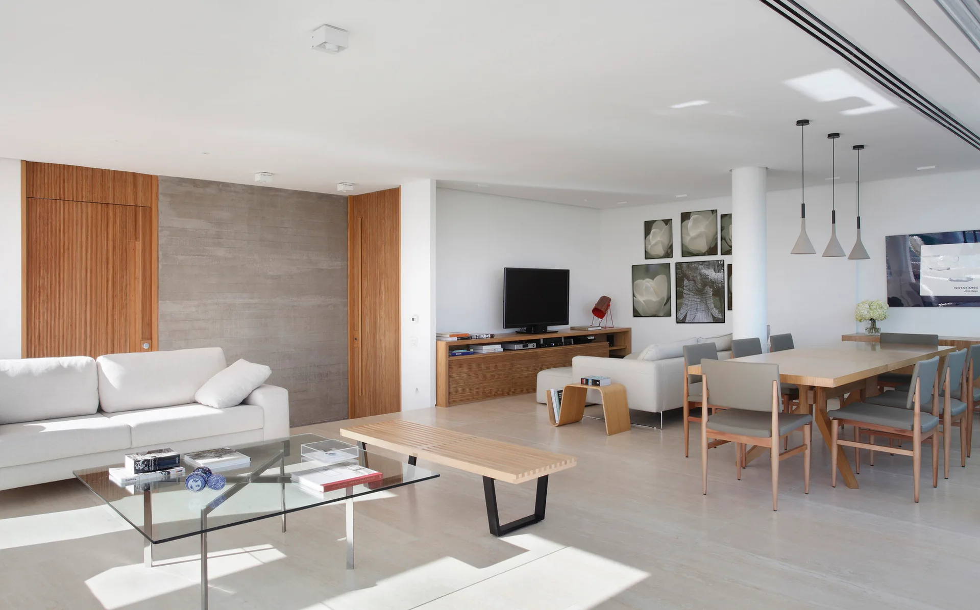 A modern open-concept living and dining area featuring a white sofa, a glass coffee table, a board-formed concrete wall, and a wooden dining table.