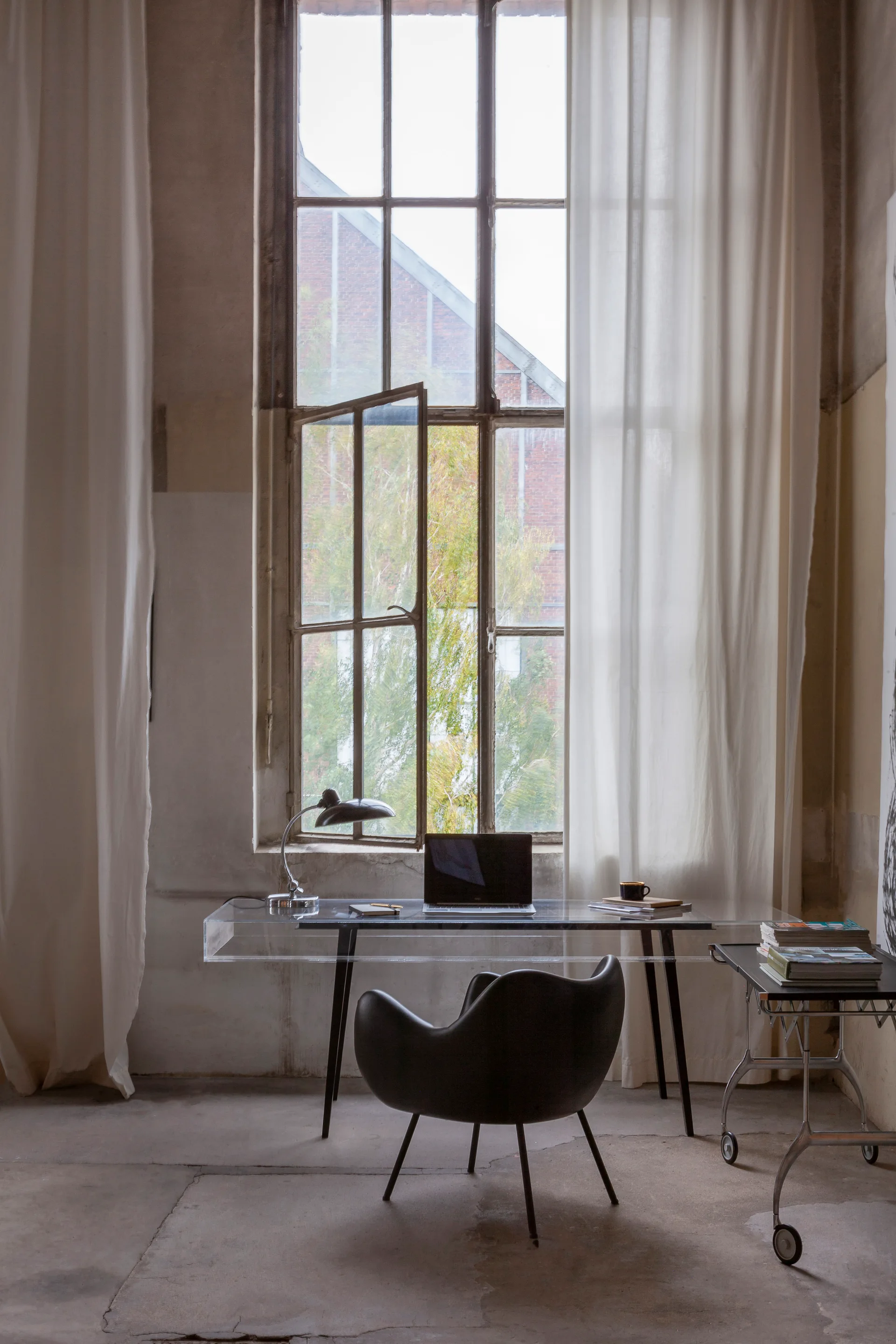 A minimalist home office featuring a transparent acrylic desk, a black molded chair, and a tall industrial window framed by white curtains.
