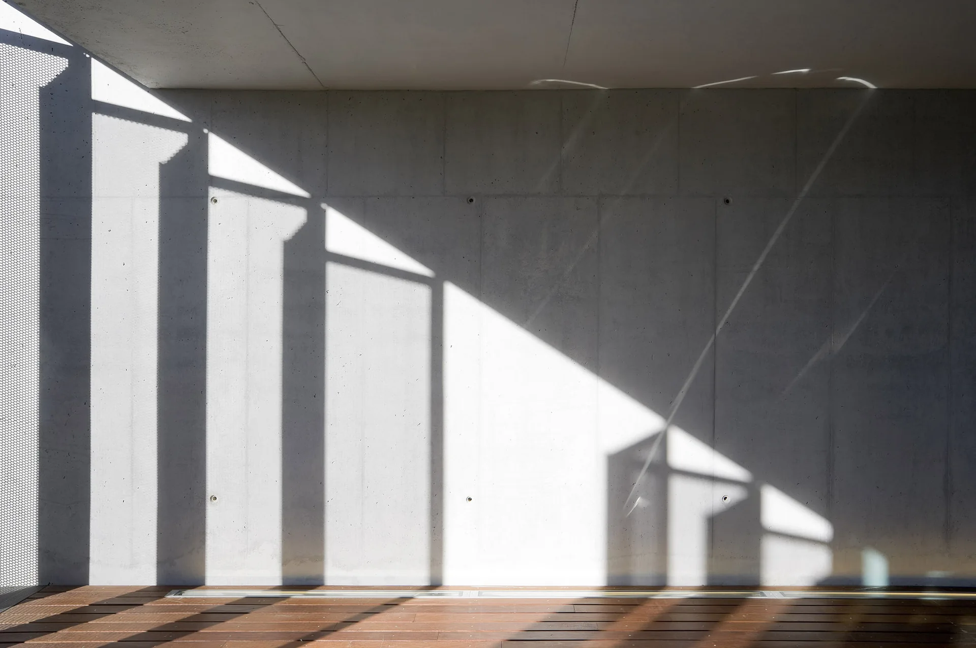 A smooth gray concrete wall features sharp geometric shadows from natural light, met by a warm wood plank floor below.