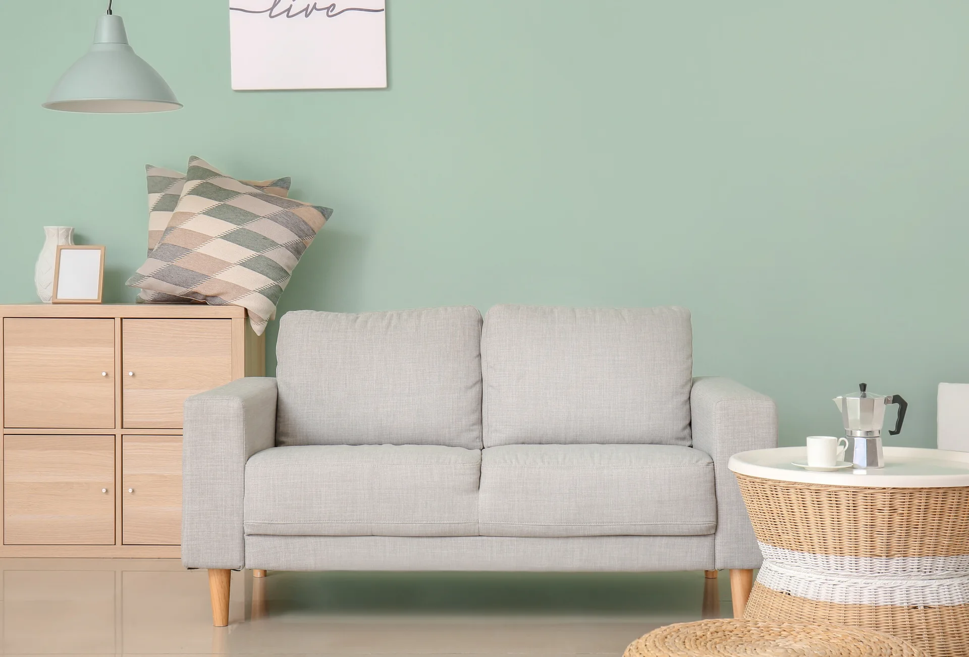 A grey fabric sofa and a round wicker coffee table sit against a soft sage green wall next to a light wood cabinet.