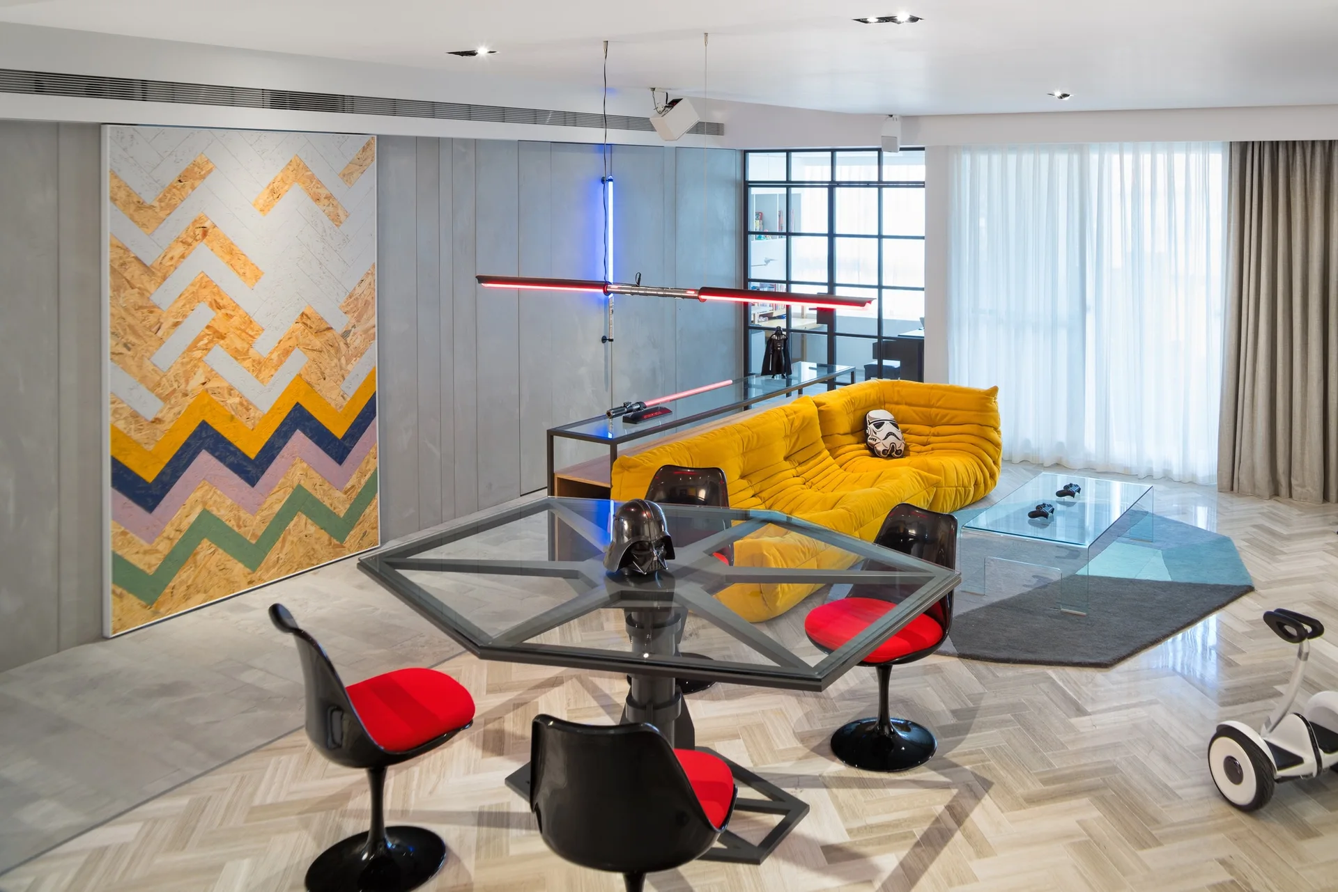 A modern open-plan living room features a yellow tufted sofa, glass dining table with black and red chairs, and grey paneled walls.