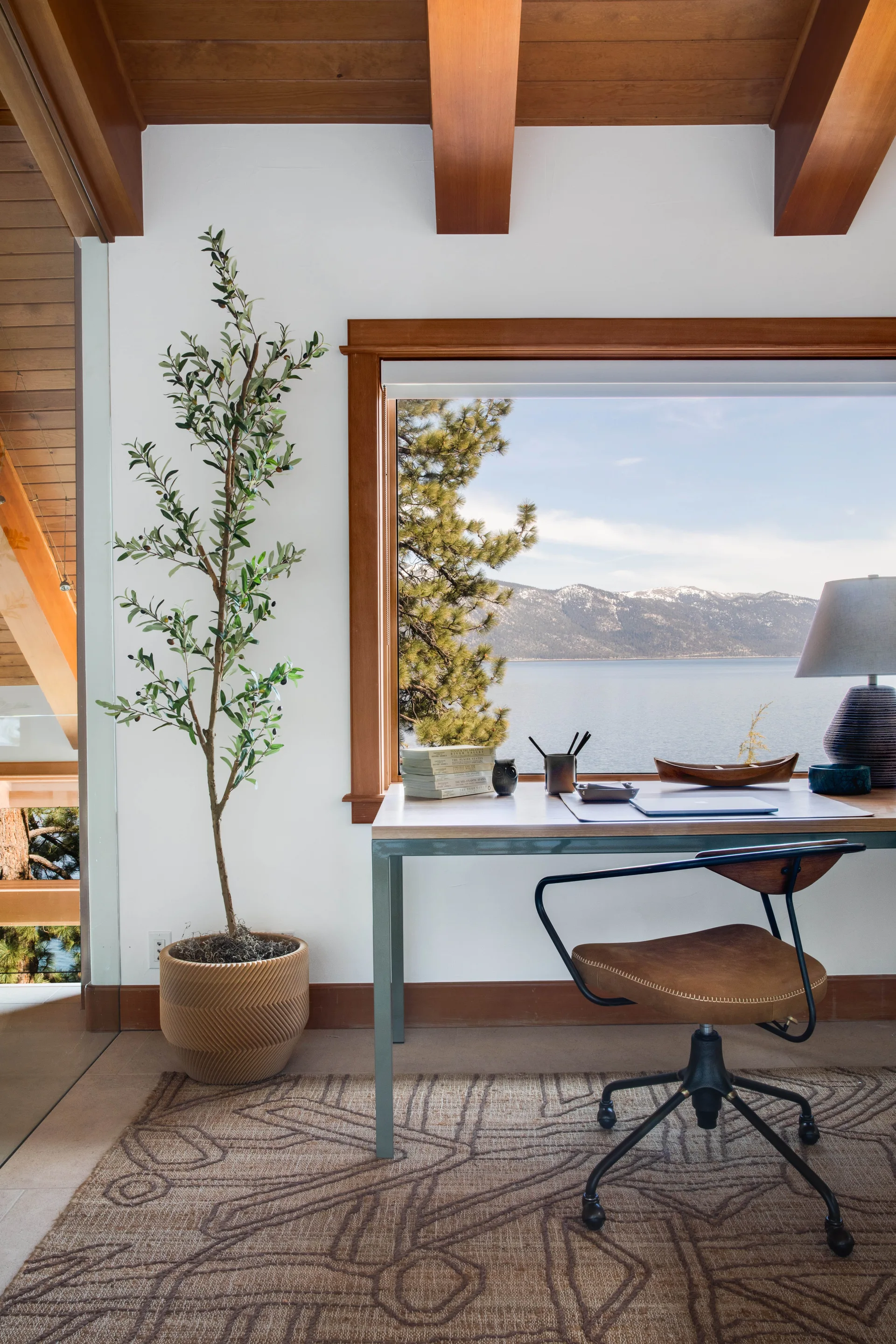 A home office features a wood-framed window with scenic lake views, an executive desk, a leather-backed chair, and exposed ceiling beams.