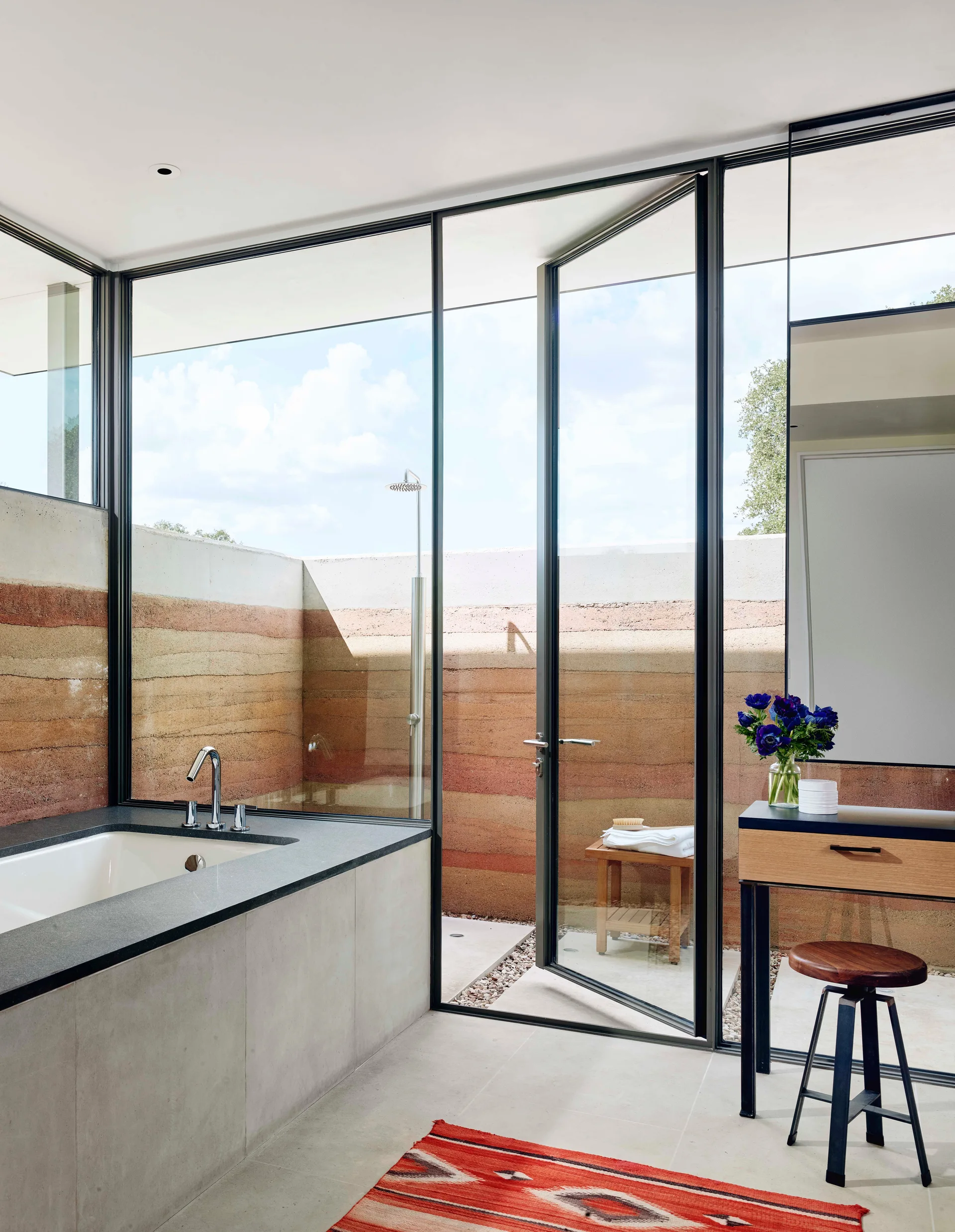 A modern bathroom with a glass door opening to an outdoor shower area enclosed by layered rammed earth walls.