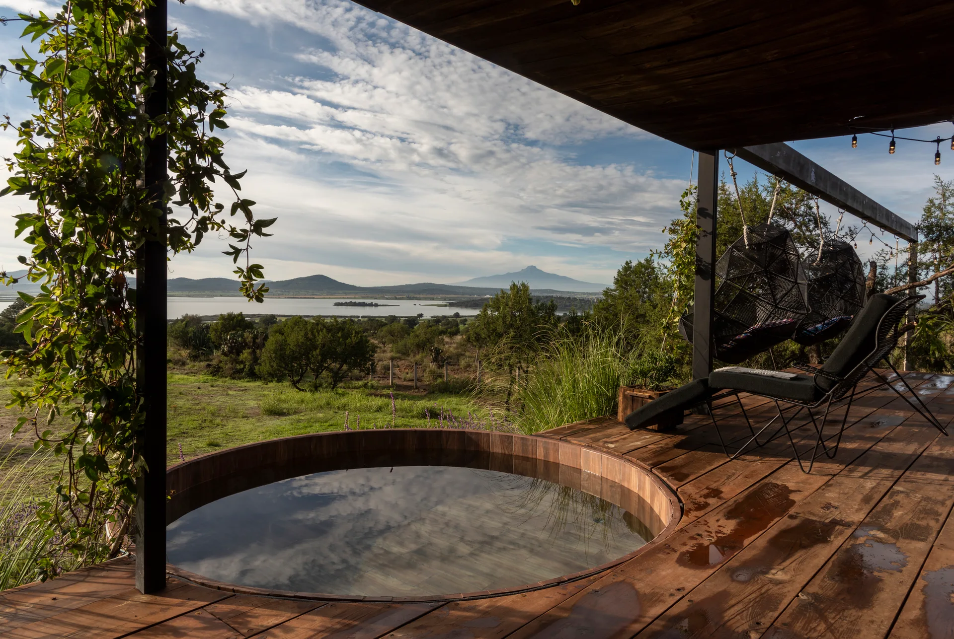 An outdoor wooden deck features a built-in circular soaking tub, black lounge chairs, and hanging pod chairs overlooking a scenic lake and mountains.
