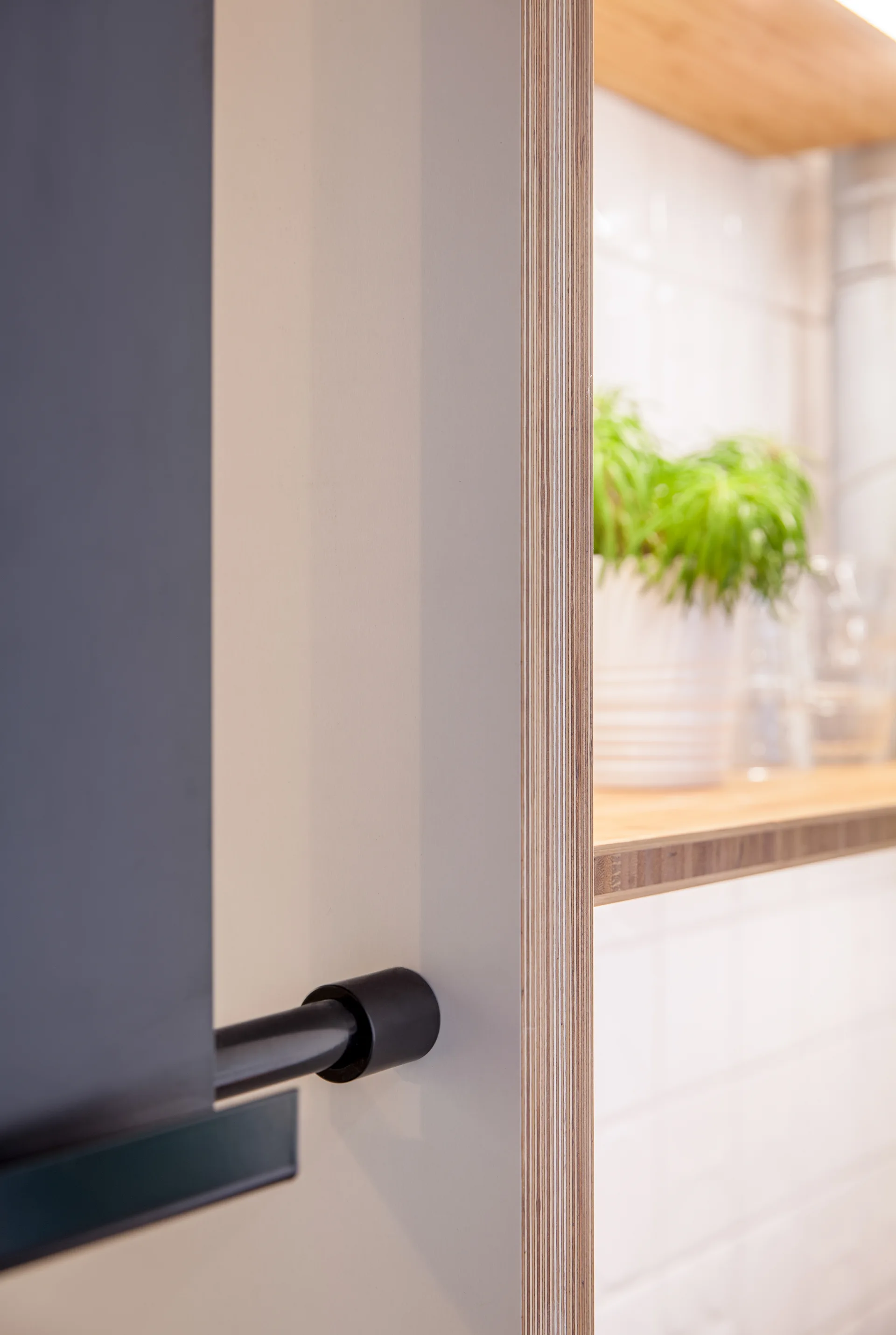 A close-up view of a matte grey cabinet with exposed plywood edges next to an open shelf holding a potted plant against a white tiled wall.