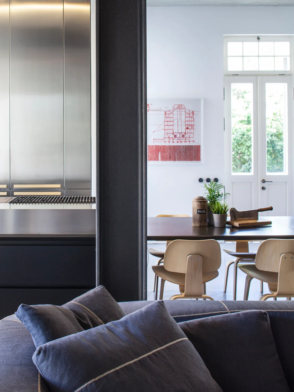 Plywood chairs surround a dining table in an open space featuring a dark structural beam and stainless steel kitchen cabinets.