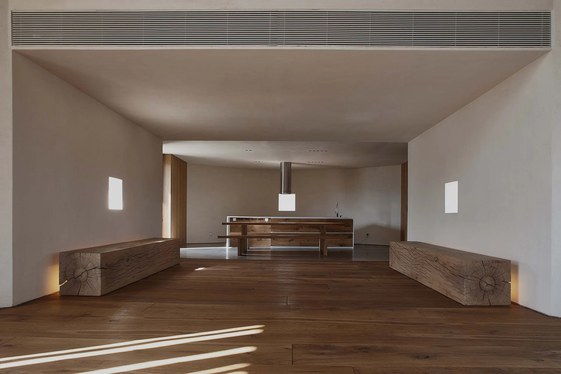 A minimalist open-plan kitchen and dining area with wide-plank wood floors, two solid log benches in the foreground, and a central kitchen island.