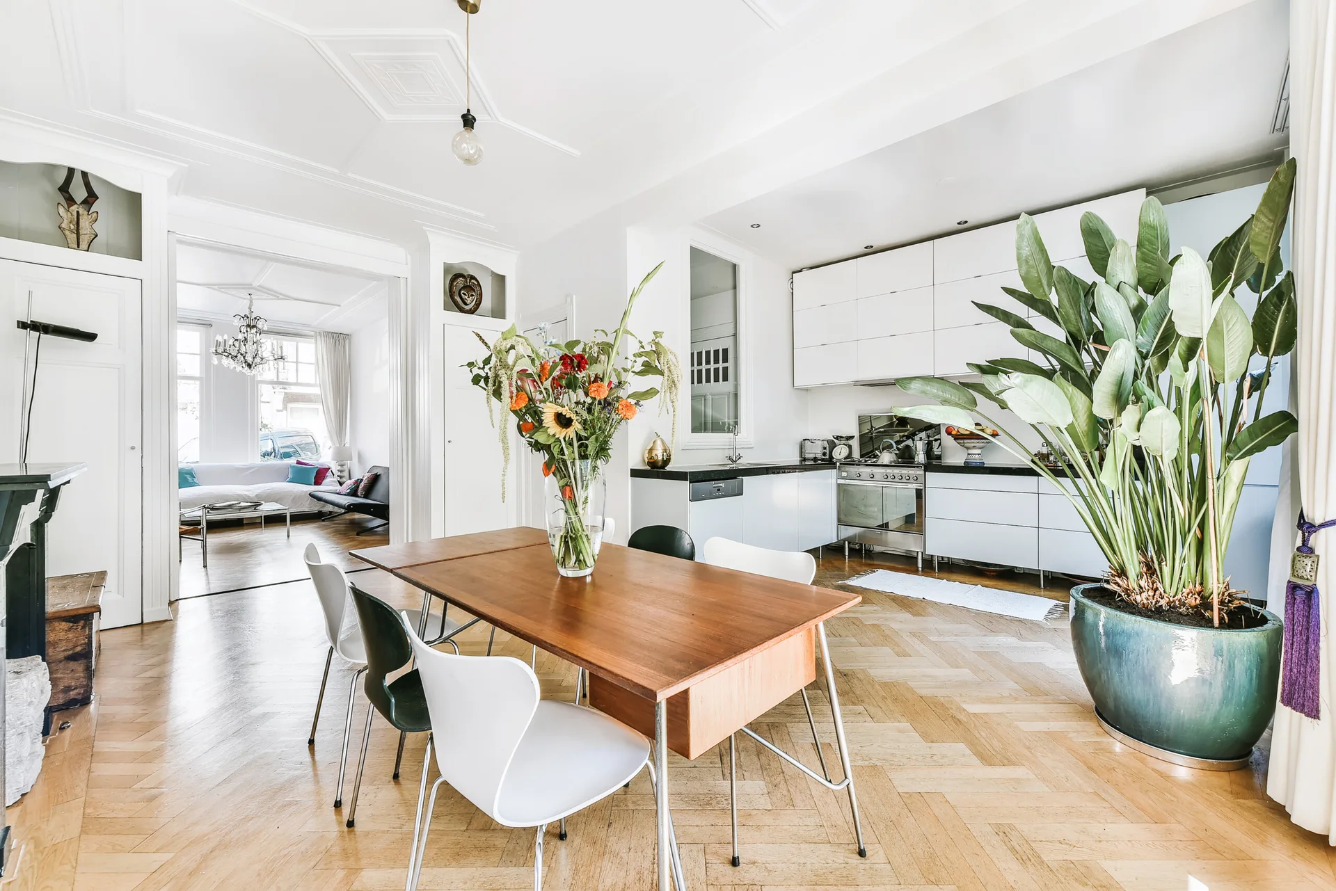 An open dining area features a wooden table with mismatched chairs, herringbone wood floors, white kitchen cabinets, and a large potted plant.