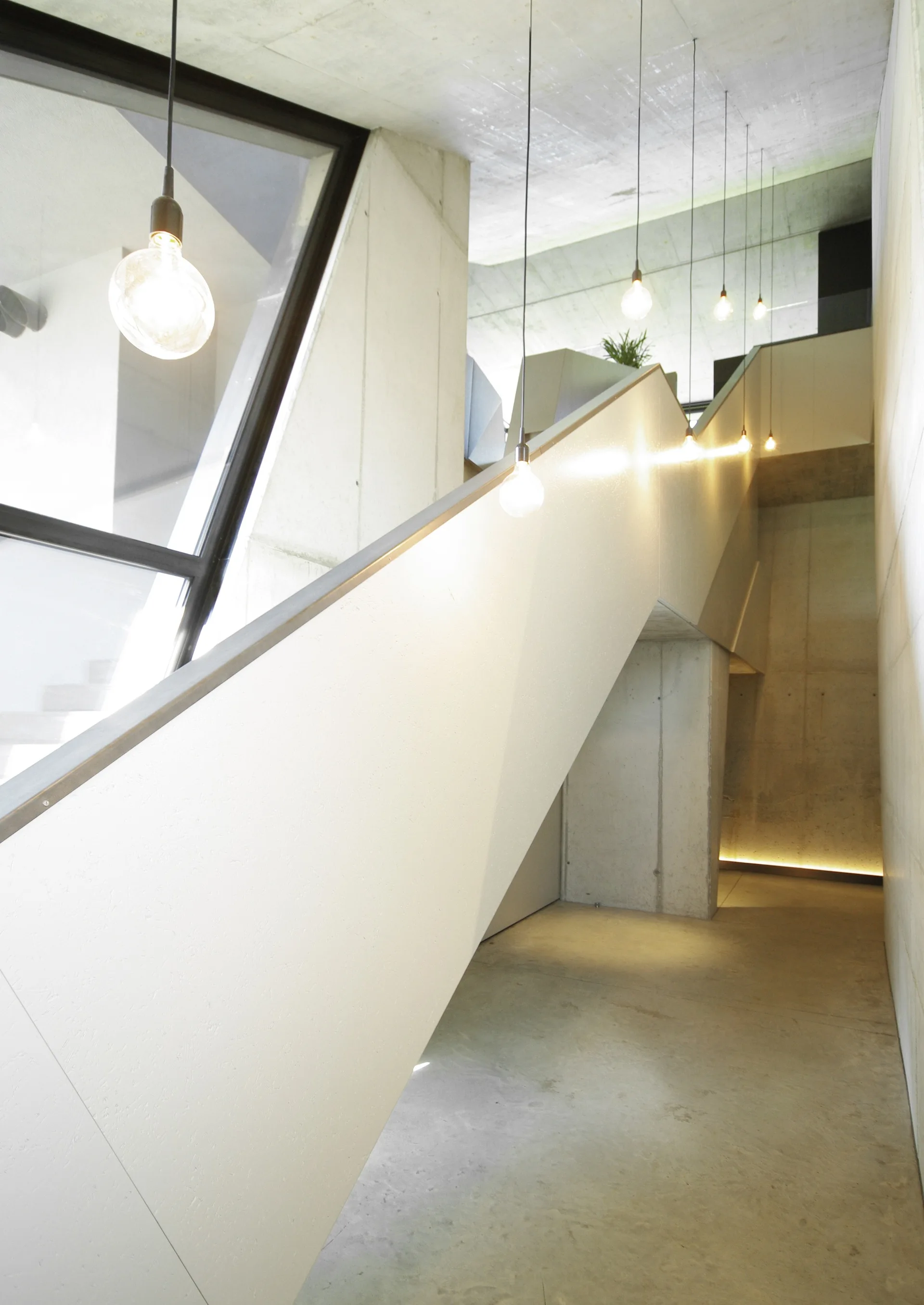 A minimalist concrete staircase rises between raw concrete walls illuminated by several hanging glass bulb pendant lights.
