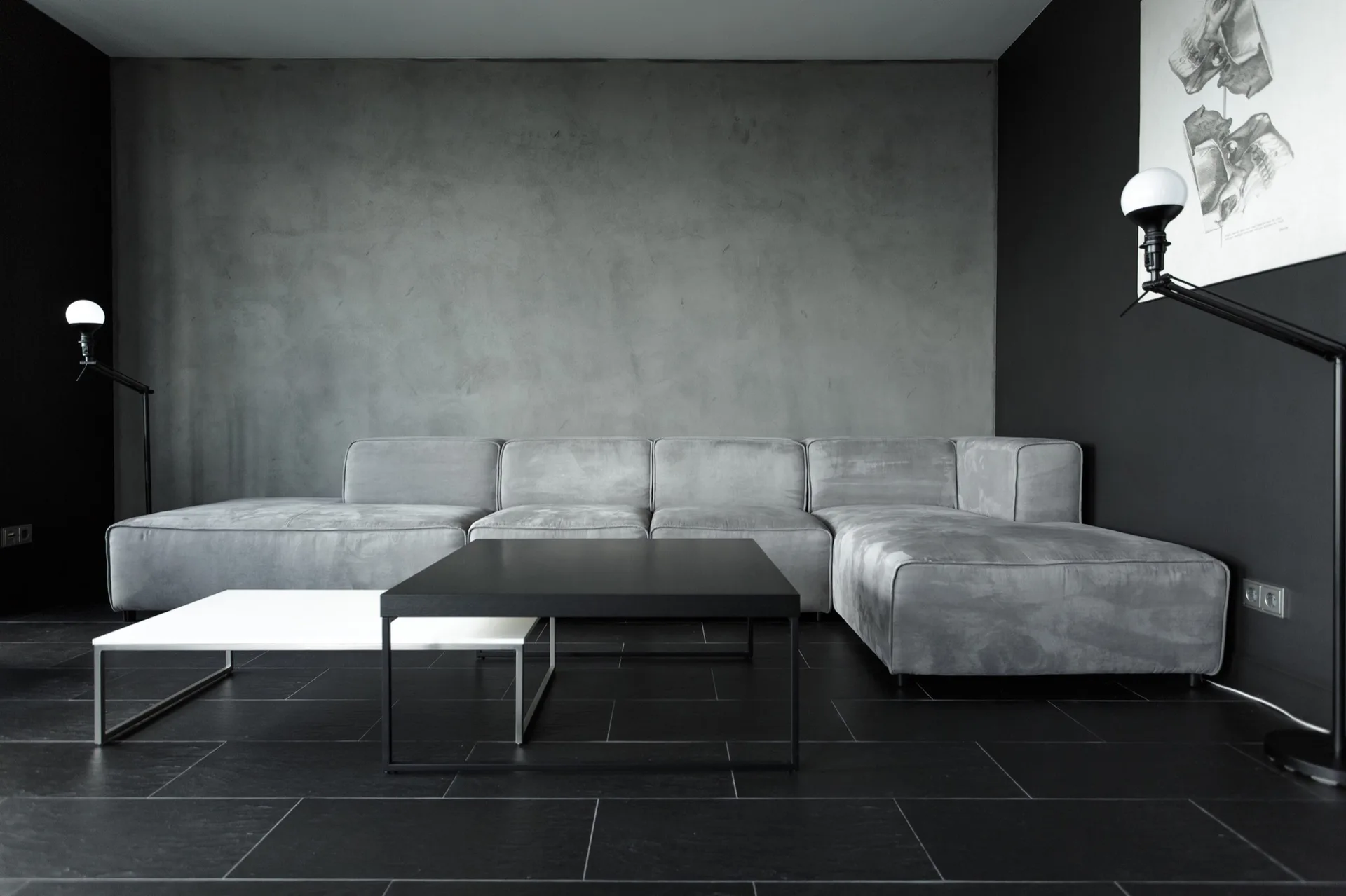A minimalist living room features a grey sectional sofa against a concrete accent wall with black floor tiles and nested coffee tables.