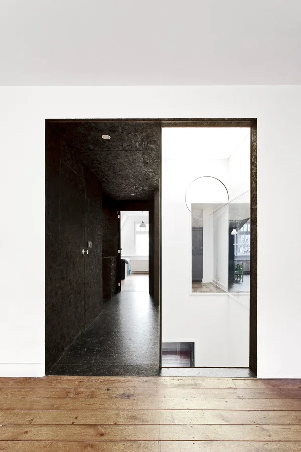 A dark, textured hallway with black walls sits adjacent to a bright, white interior room, separated by a glass panel and set above wood flooring.