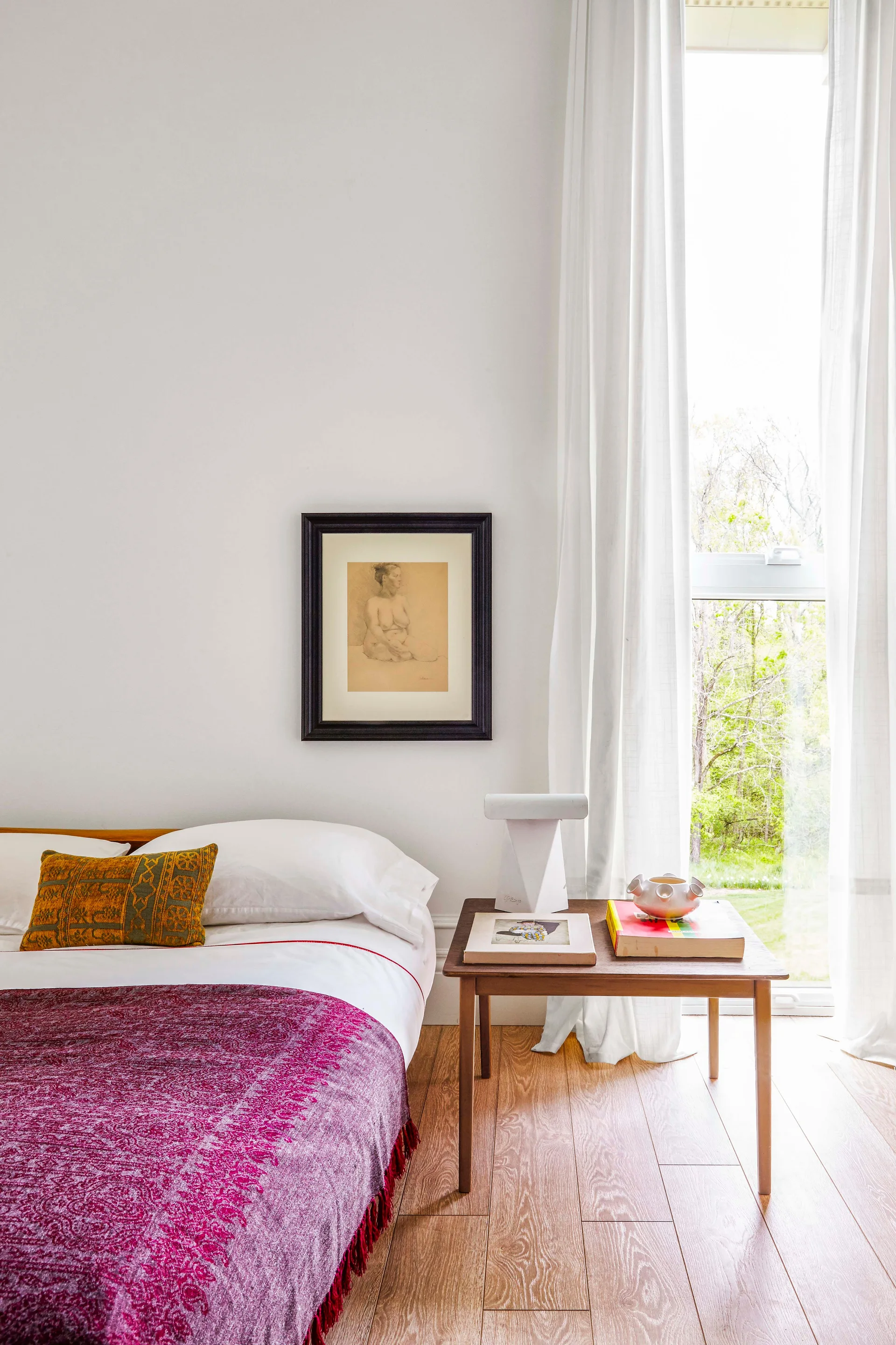 A bed with white linens and a magenta throw sits next to a wooden side table with books and a lamp beneath a tall window with sheer curtains.