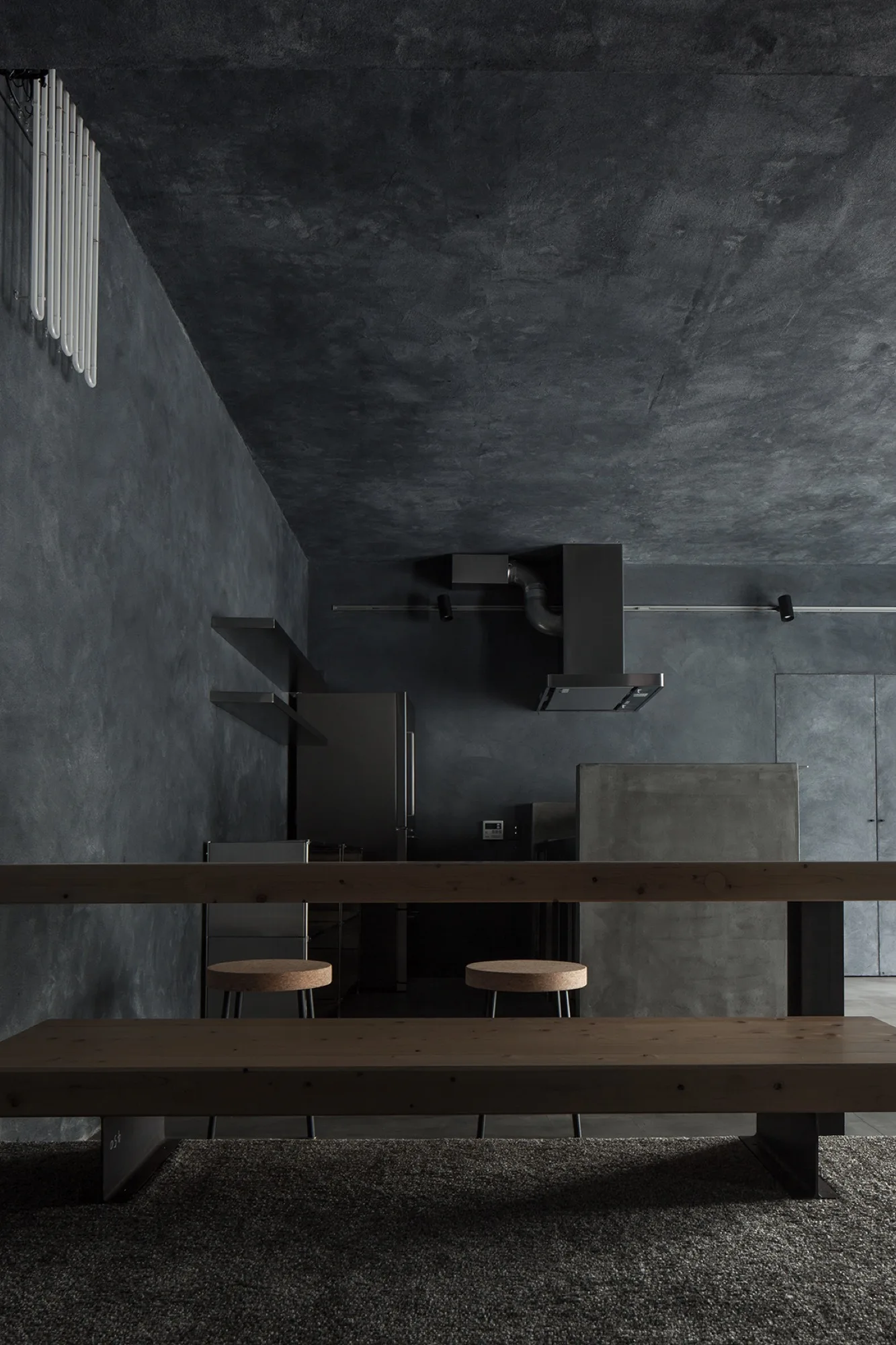 A minimalist charcoal kitchen features textured plaster walls, a concrete island, and a long wooden dining table with cork-topped stools in the foreground.