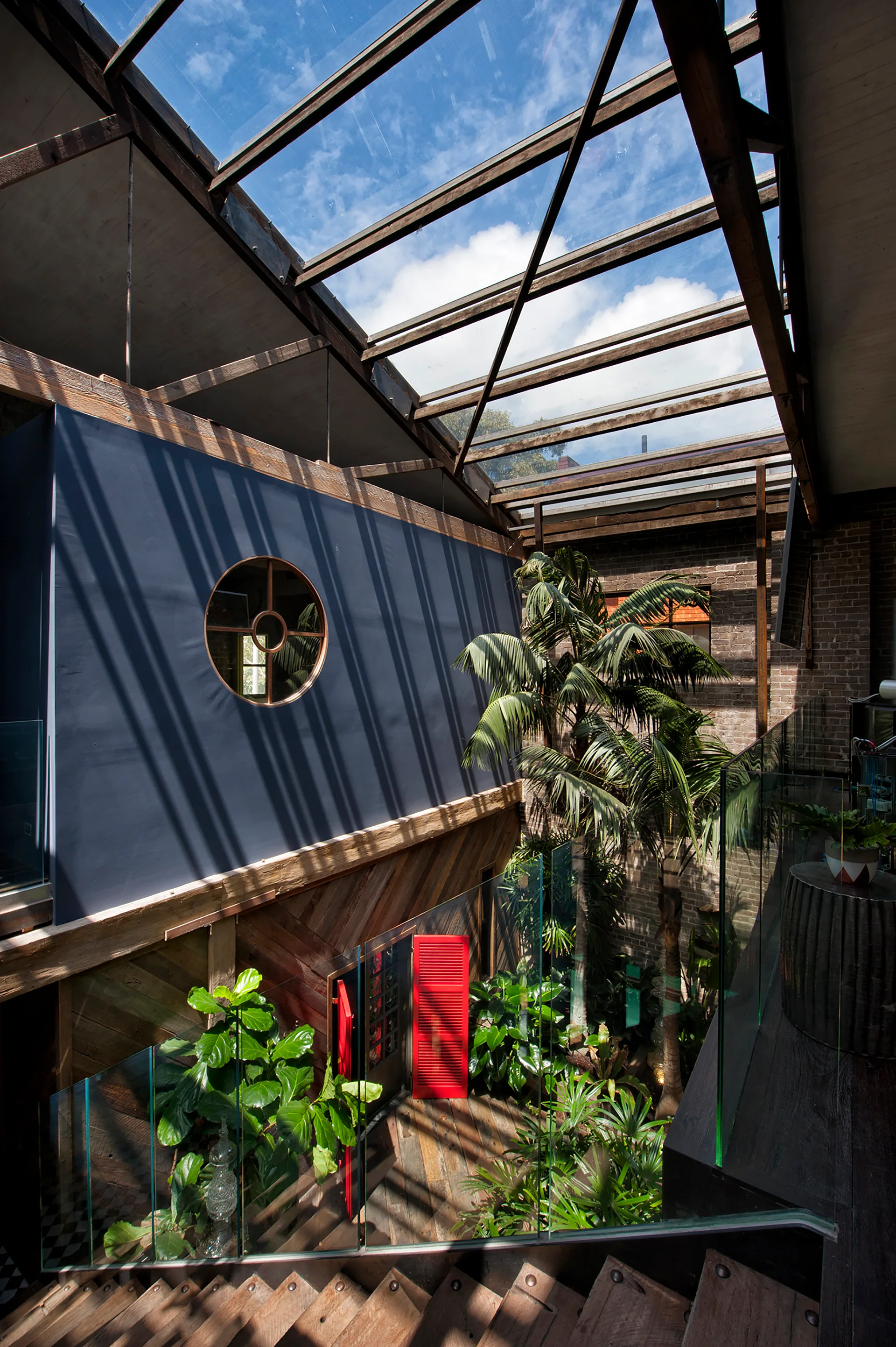 Looking down a wooden staircase into an indoor courtyard with a glass skylight, navy blue walls, lush green plants, and a red louvered door.