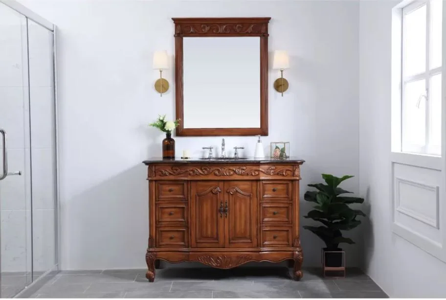 An ornate brown wood vanity with a dark countertop sits against a white wall beneath a matching wood-framed mirror and brass sconces.