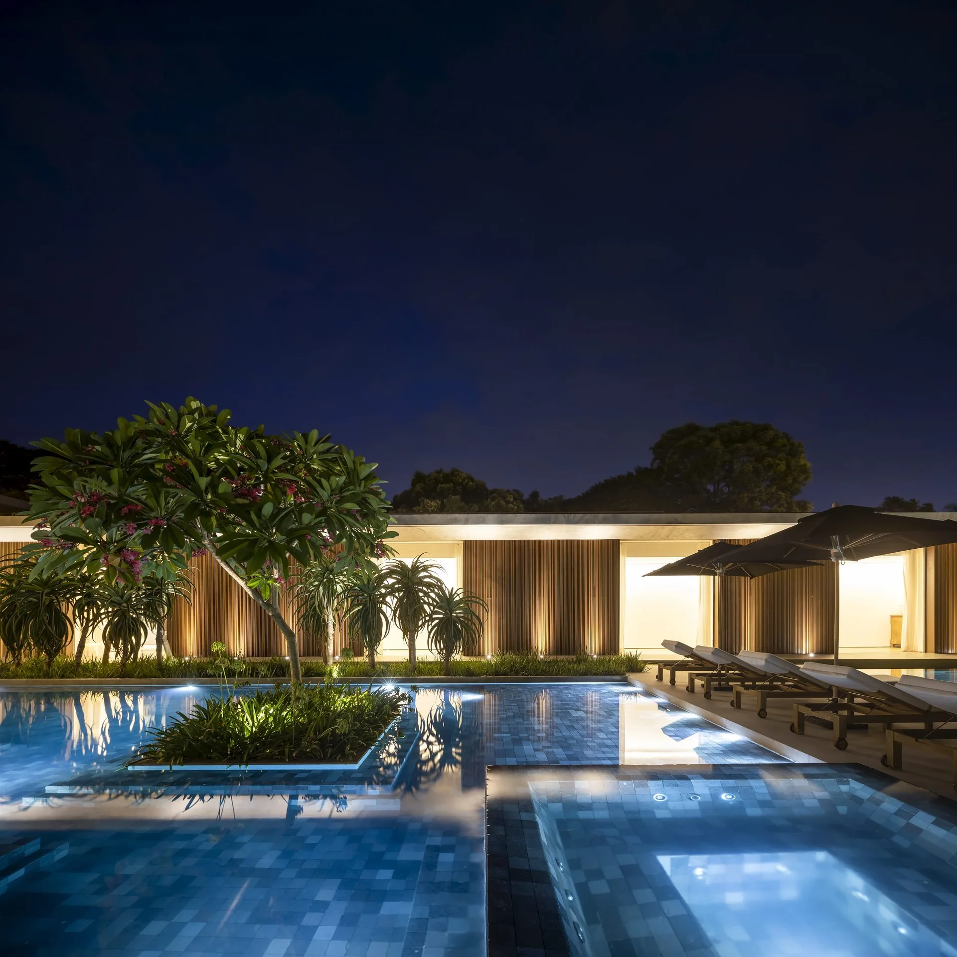 An illuminated outdoor swimming pool at night features a central planting island, lounge chairs with umbrellas, and a modern slatted wood building facade.