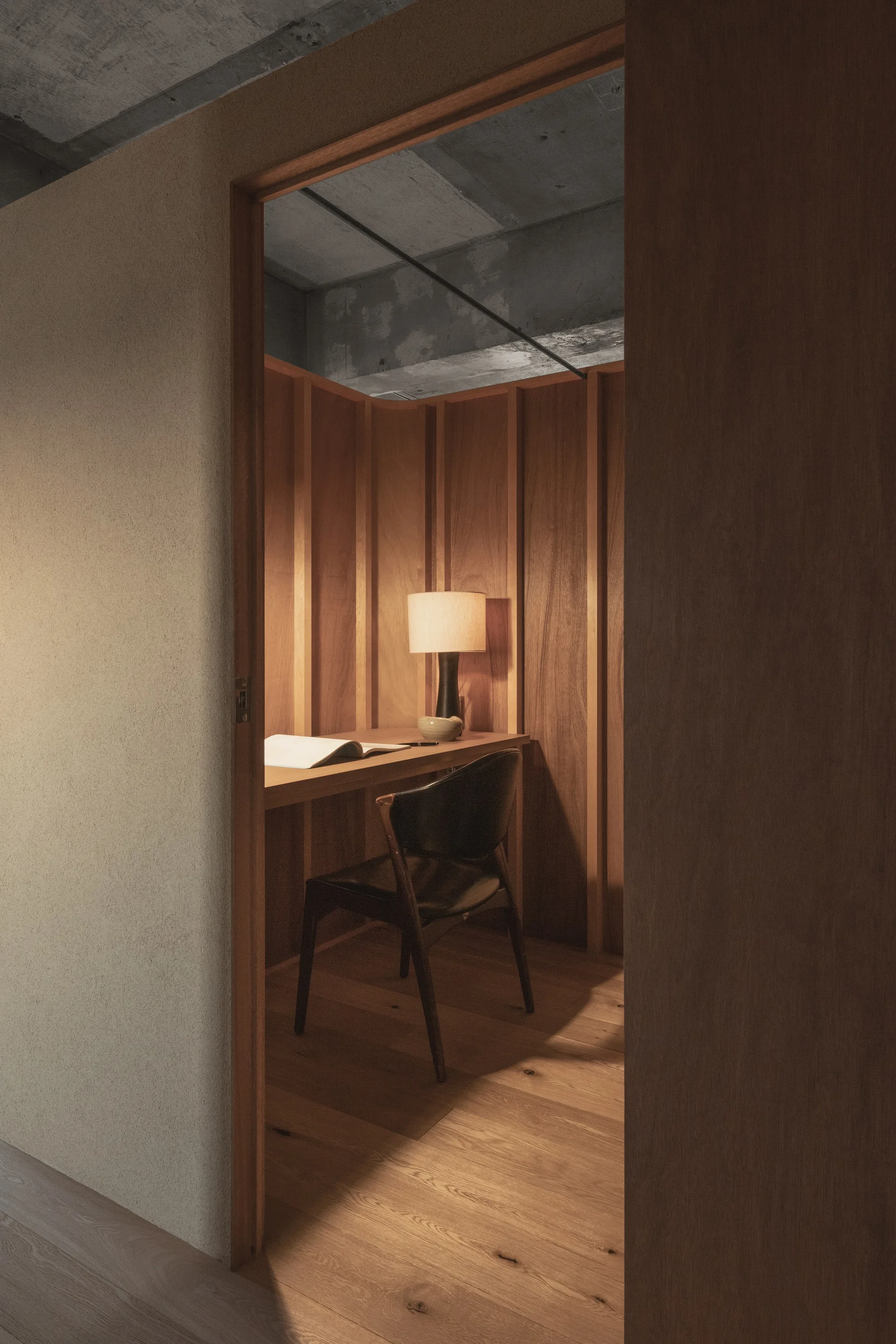 View through a doorway into a small workspace with vertical wood paneling, a built-in wooden desk, a black chair, and an exposed concrete ceiling.