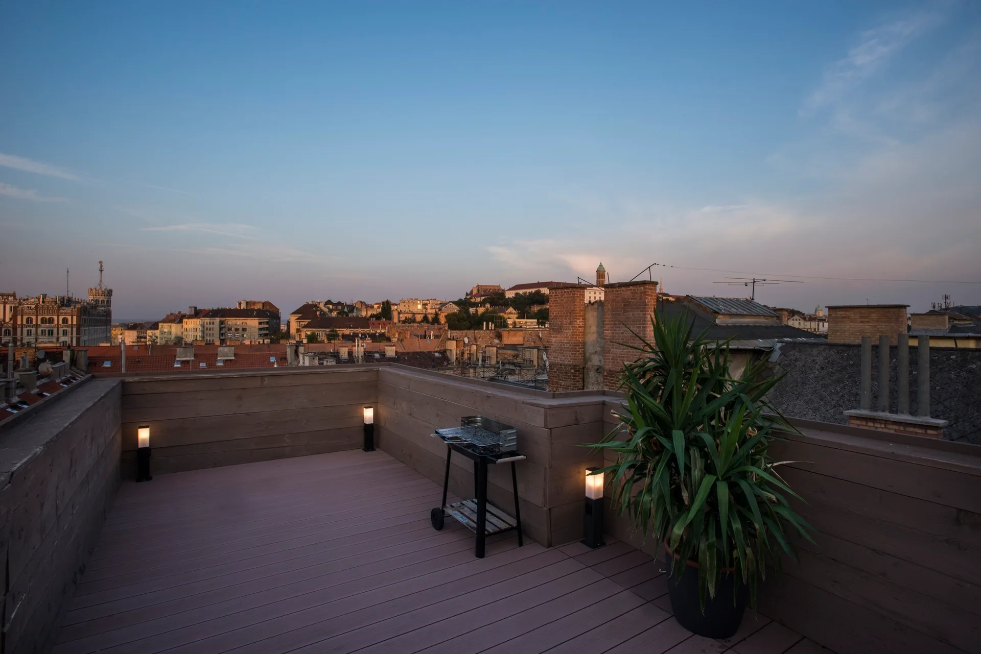 A rooftop wooden deck features a portable charcoal grill, a large potted plant, and warm post lights against a background city skyline.
