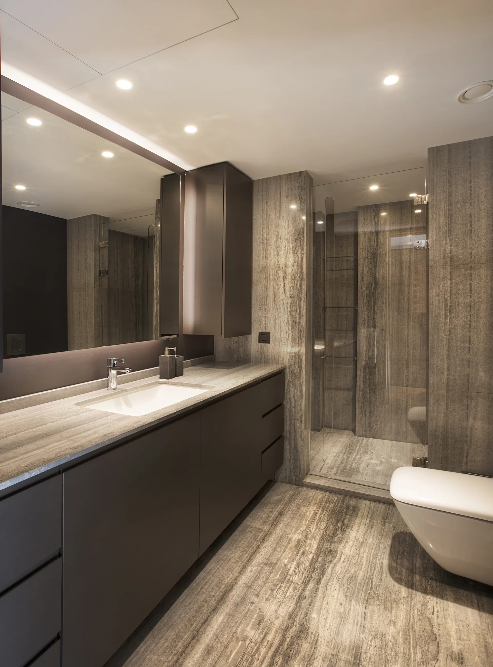 A modern bathroom features grey veined stone tile walls and floors, a dark brown floating vanity with a large mirror, and a wall-hung toilet.