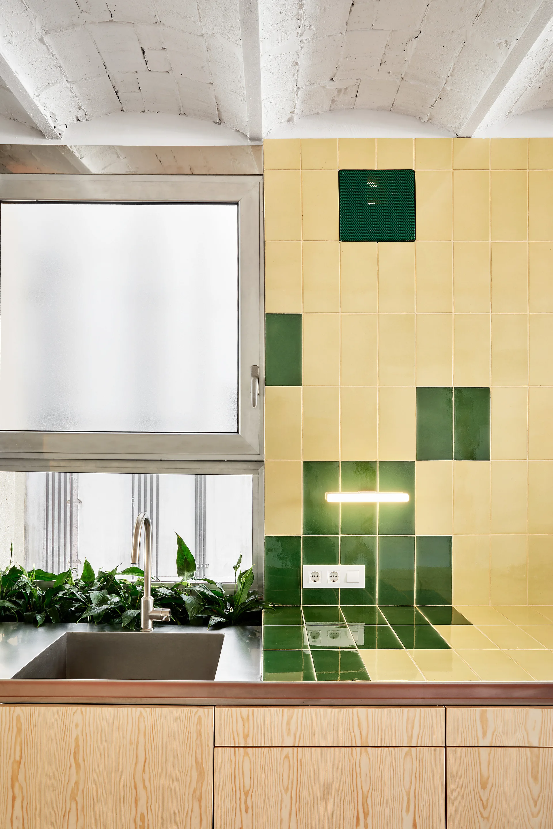 A modern kitchen sink area features a yellow and green tiled backsplash, light wood lower cabinets, and a white vaulted ceiling.