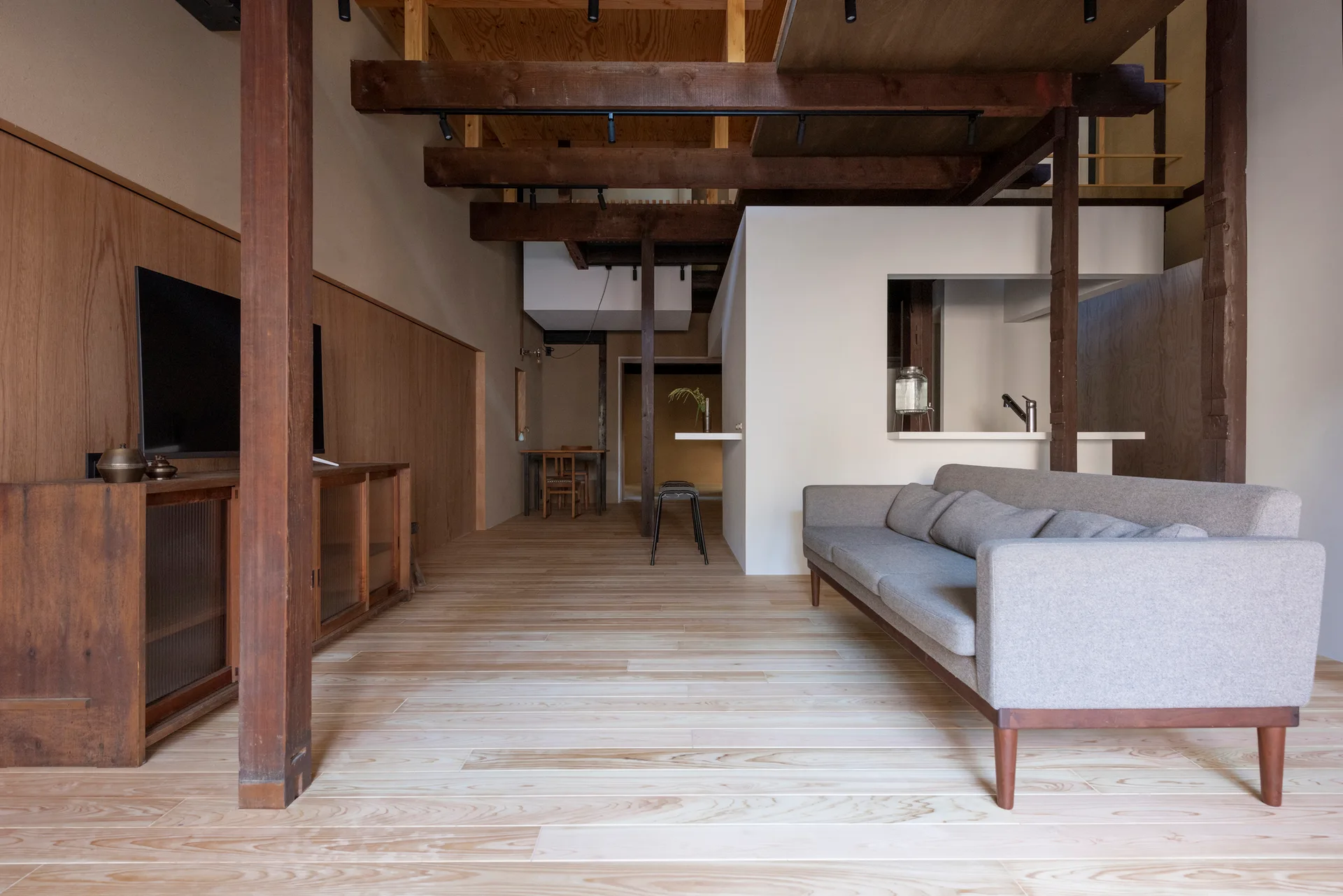 A minimalist open-concept living room featuring light wood flooring, exposed dark timber ceiling beams, and a modern grey sofa.