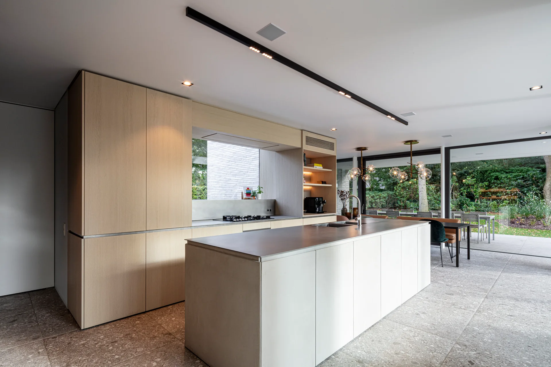 A modern kitchen featuring tall light wood cabinets, a large central island with a grey top, and sliding glass doors opening to a garden.