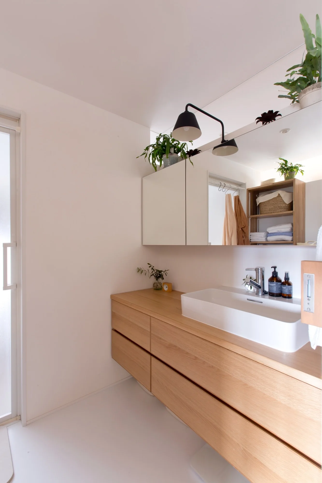 A light wood floating bathroom vanity supports a white vessel sink beneath a large mirrored cabinet and a black double-headed wall light.