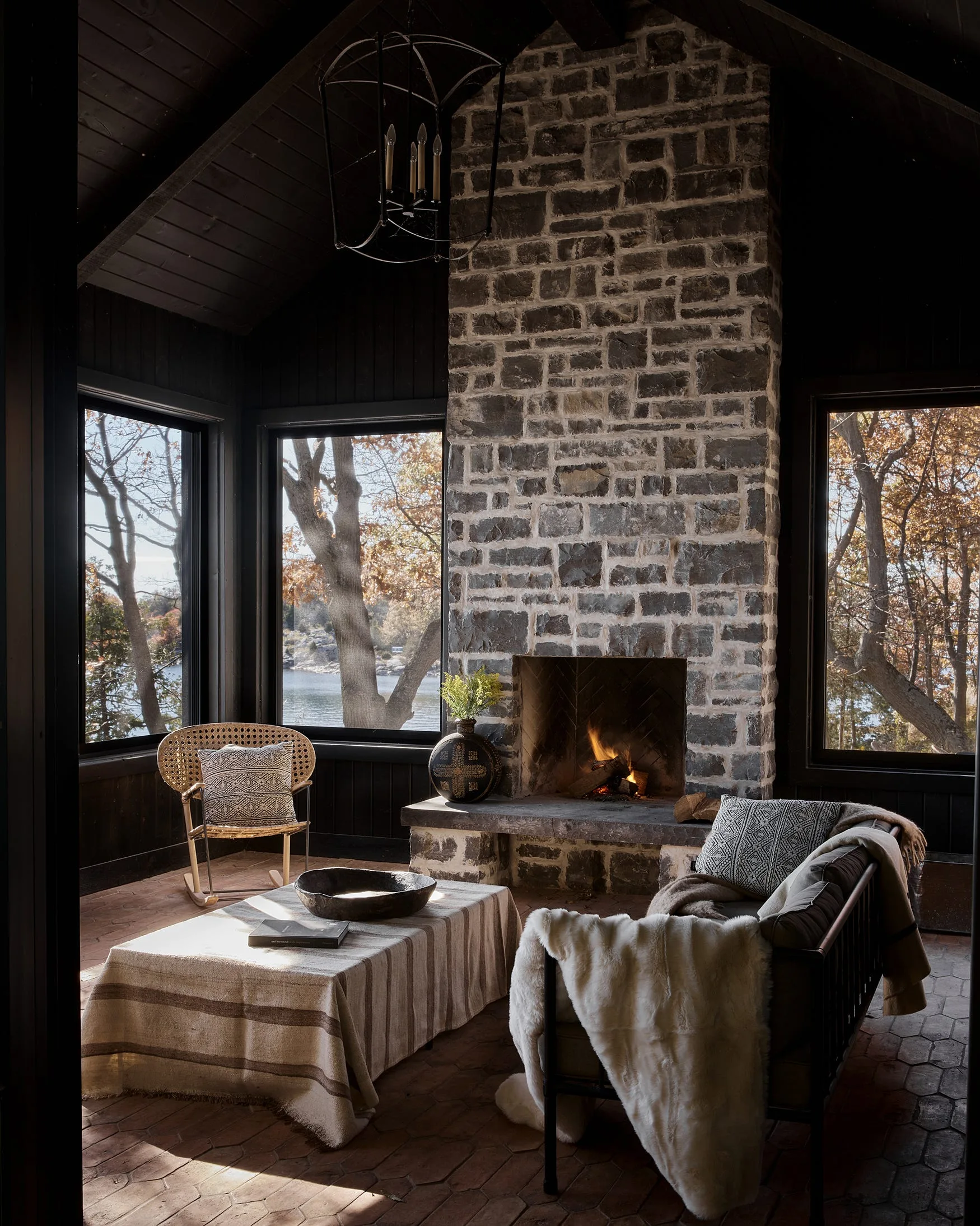 A rustic sunroom features a tall stacked stone fireplace, dark wood paneled walls, a rattan chair, and large windows looking out to a lake.
