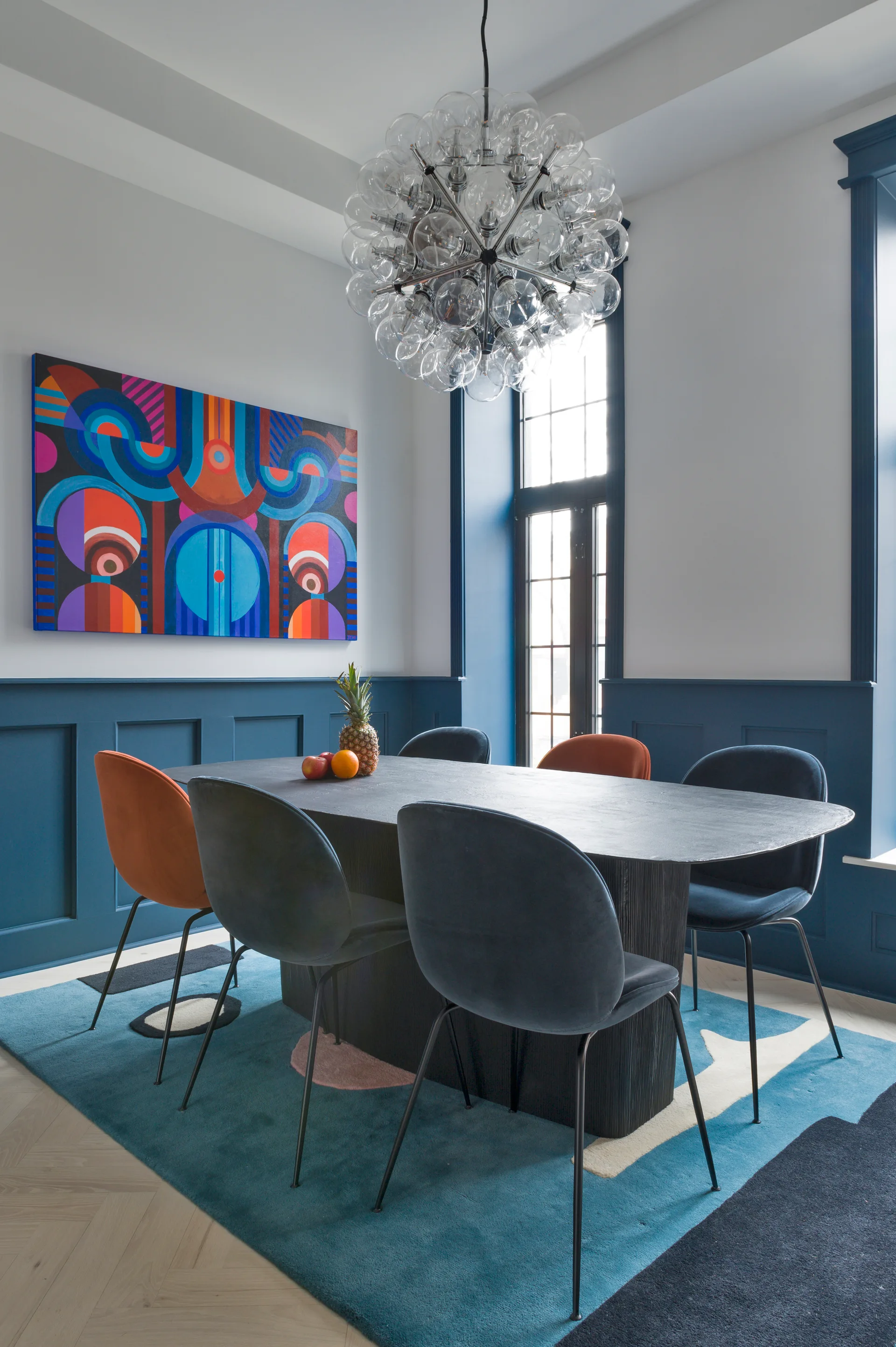 A modern dining room with a black oval table, blue and orange velvet chairs, a glass bubble chandelier, and dark blue wainscoting on the walls.