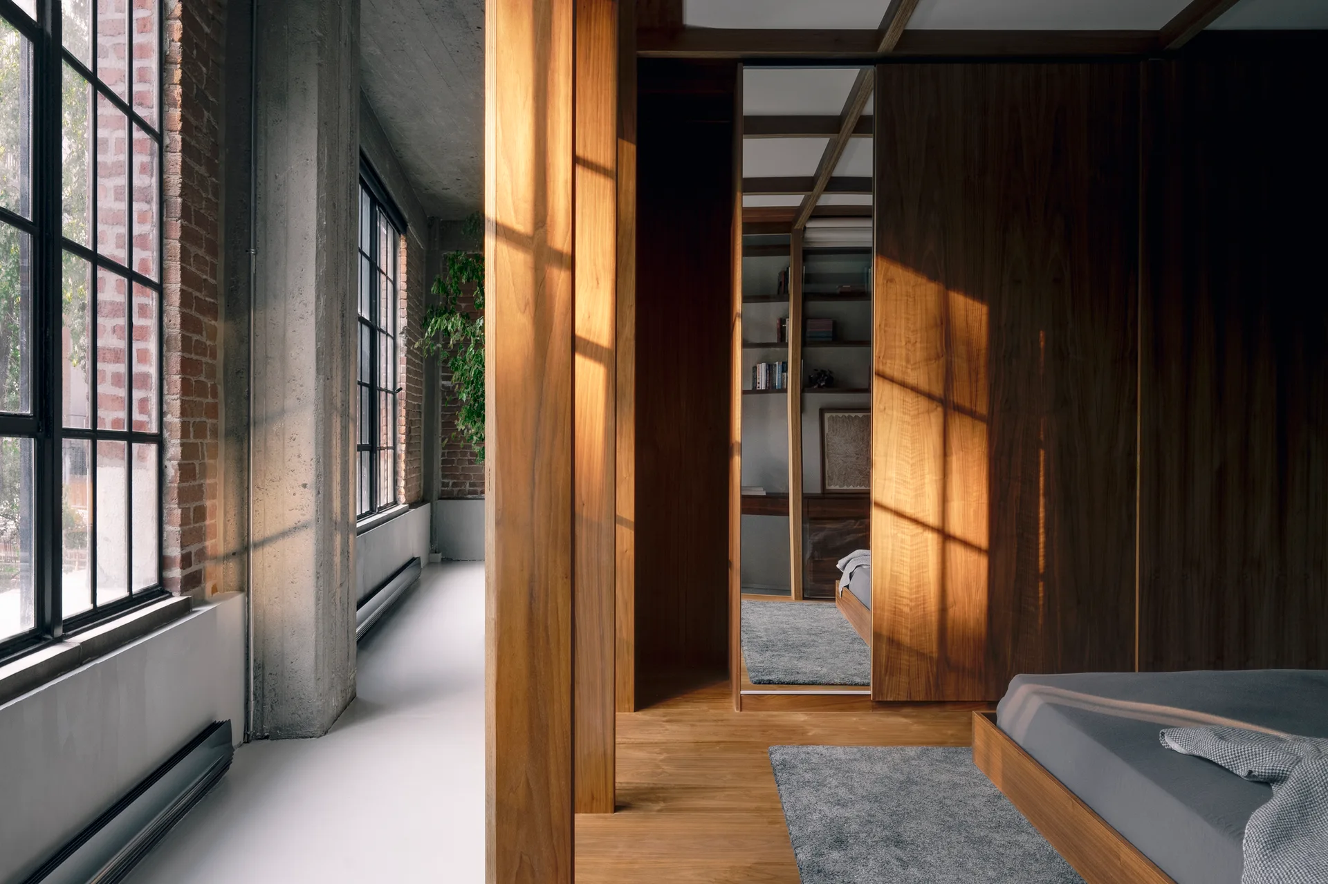 An industrial loft bedroom featuring a warm wood-paneled wardrobe, an exposed concrete column, and large black-framed windows.