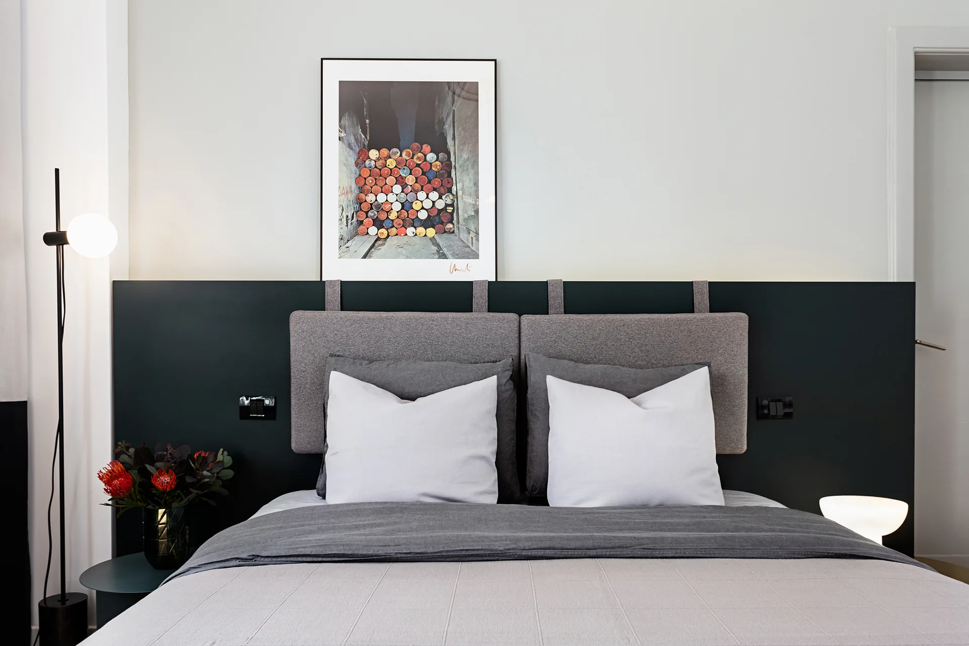 A minimalist bedroom featuring a bed with grey pillows, a dark grey horizontal wall panel with hanging cushions, a globe floor lamp, and framed art.