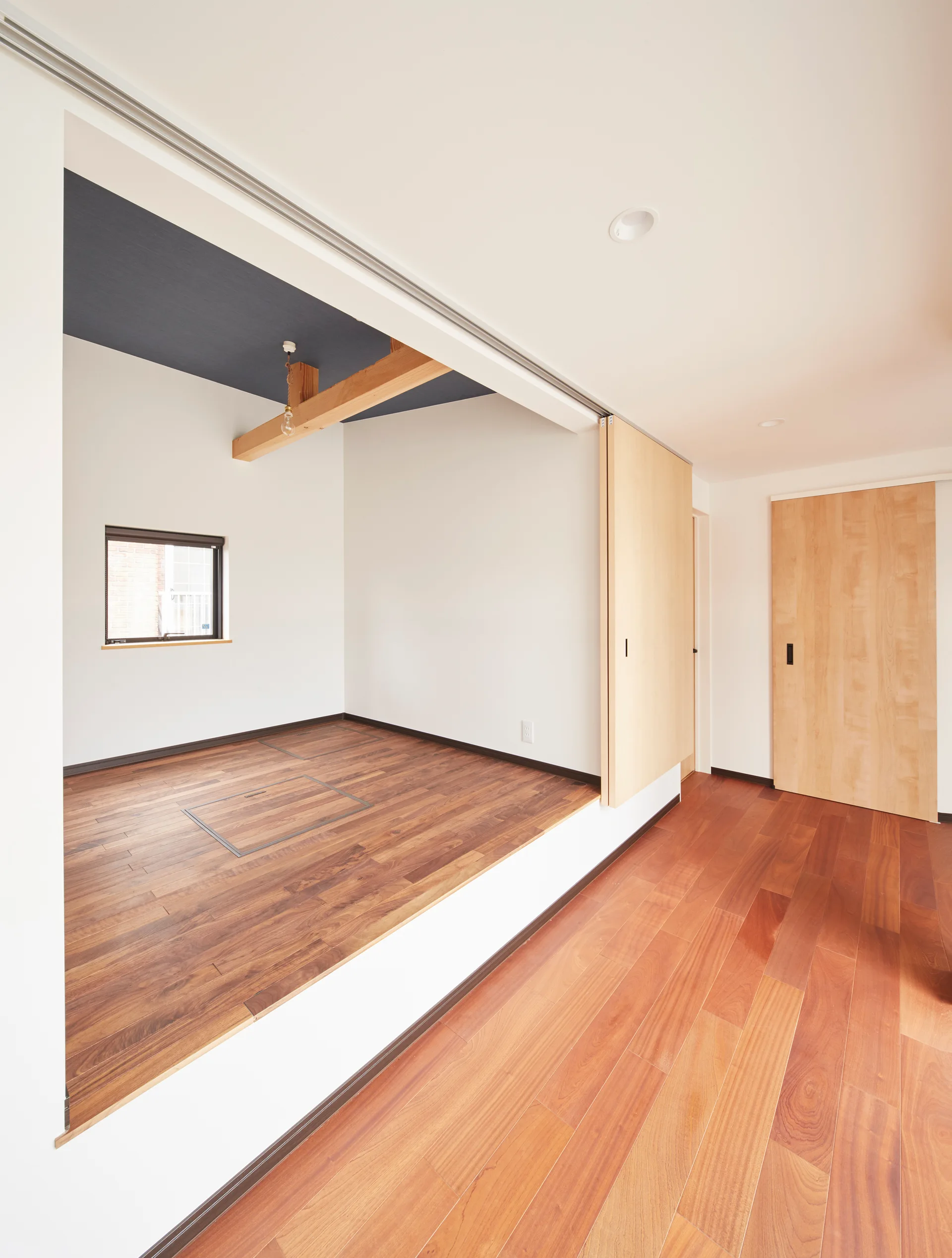 A minimalist room features an elevated wooden platform with a small window, an exposed beam ceiling, and a light wood sliding partition door.