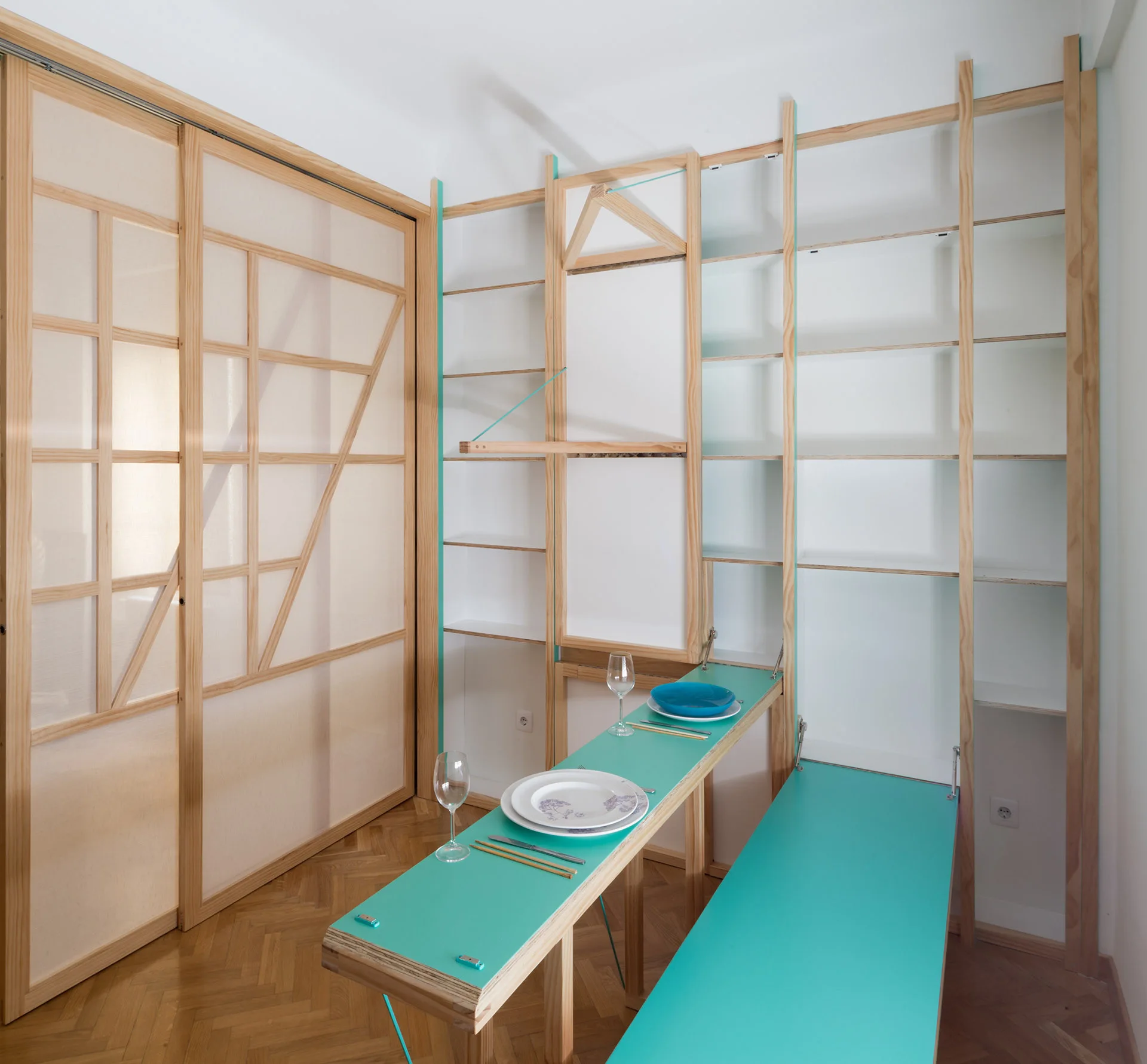 A small dining setup featuring a fold-out teal table and bench integrated into a light wood shelving unit next to a sliding Shoji-style door.