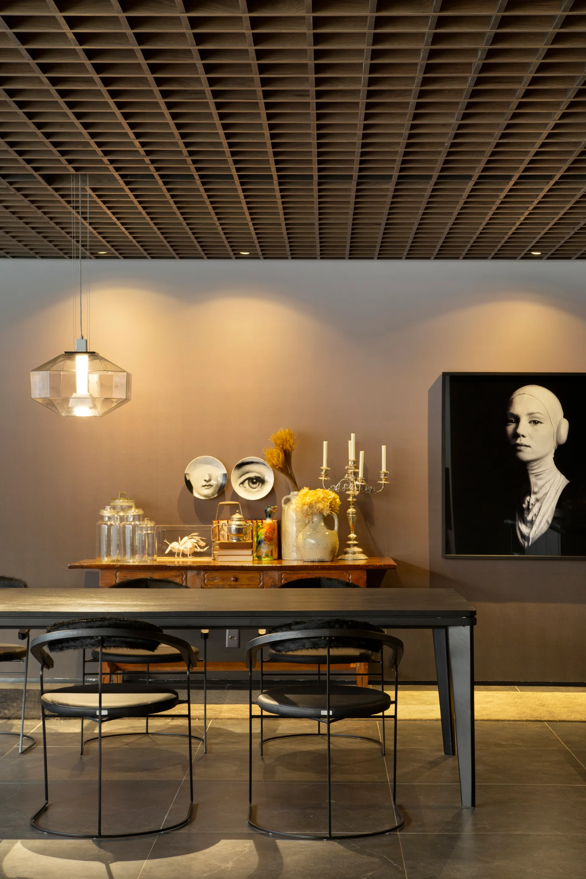 A dark dining room features a wood grid ceiling, black table and chairs, a wooden sideboard with decor, and a large black and white portrait.